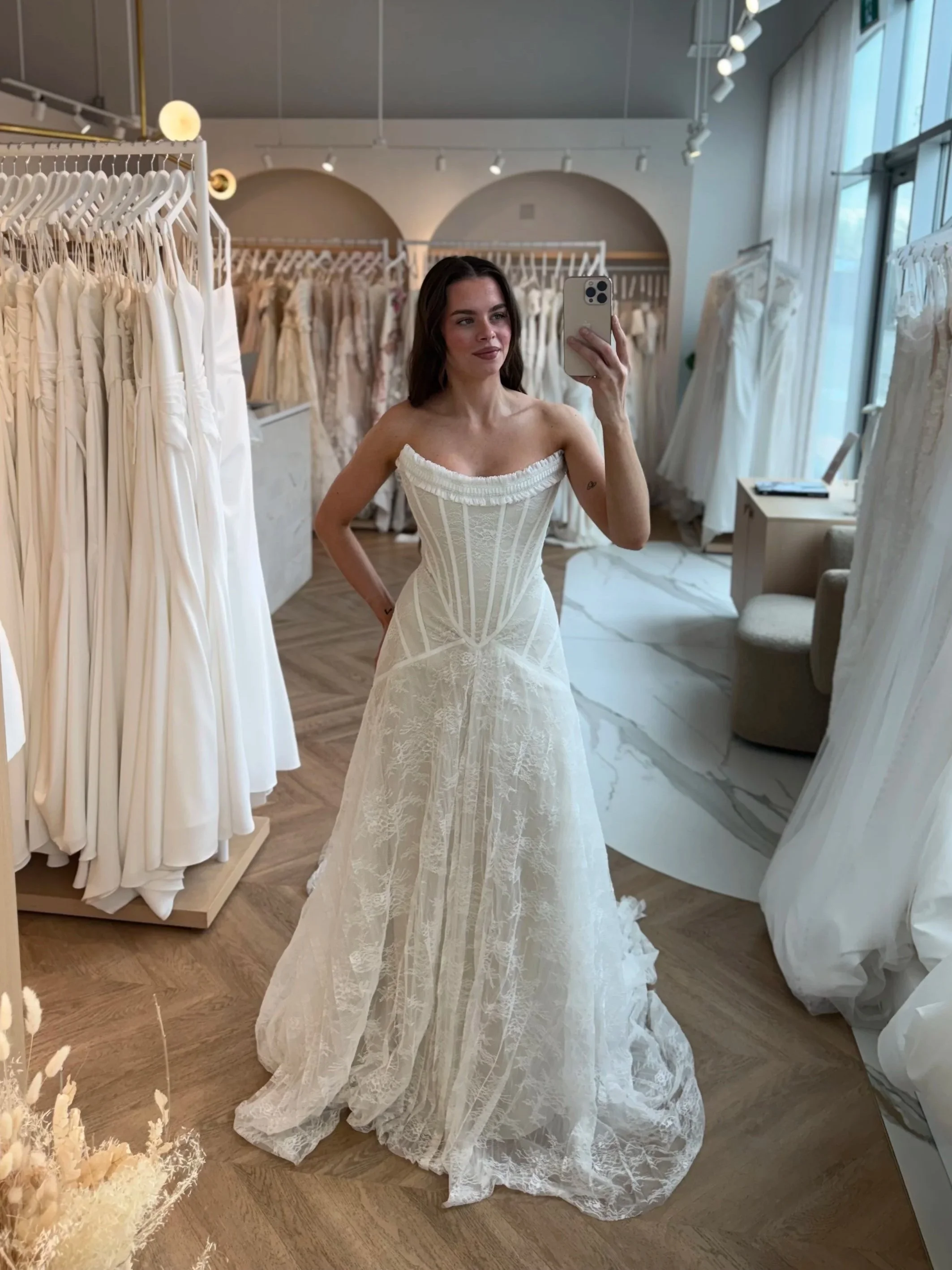 A stunning bride taking a selfie in Rue De Seine Macbeth at The Bridal Boutique. With beautiful dresses in the background. The dress is a Basque waist with Chantilly lace and a ruffle sharp scoop neckline