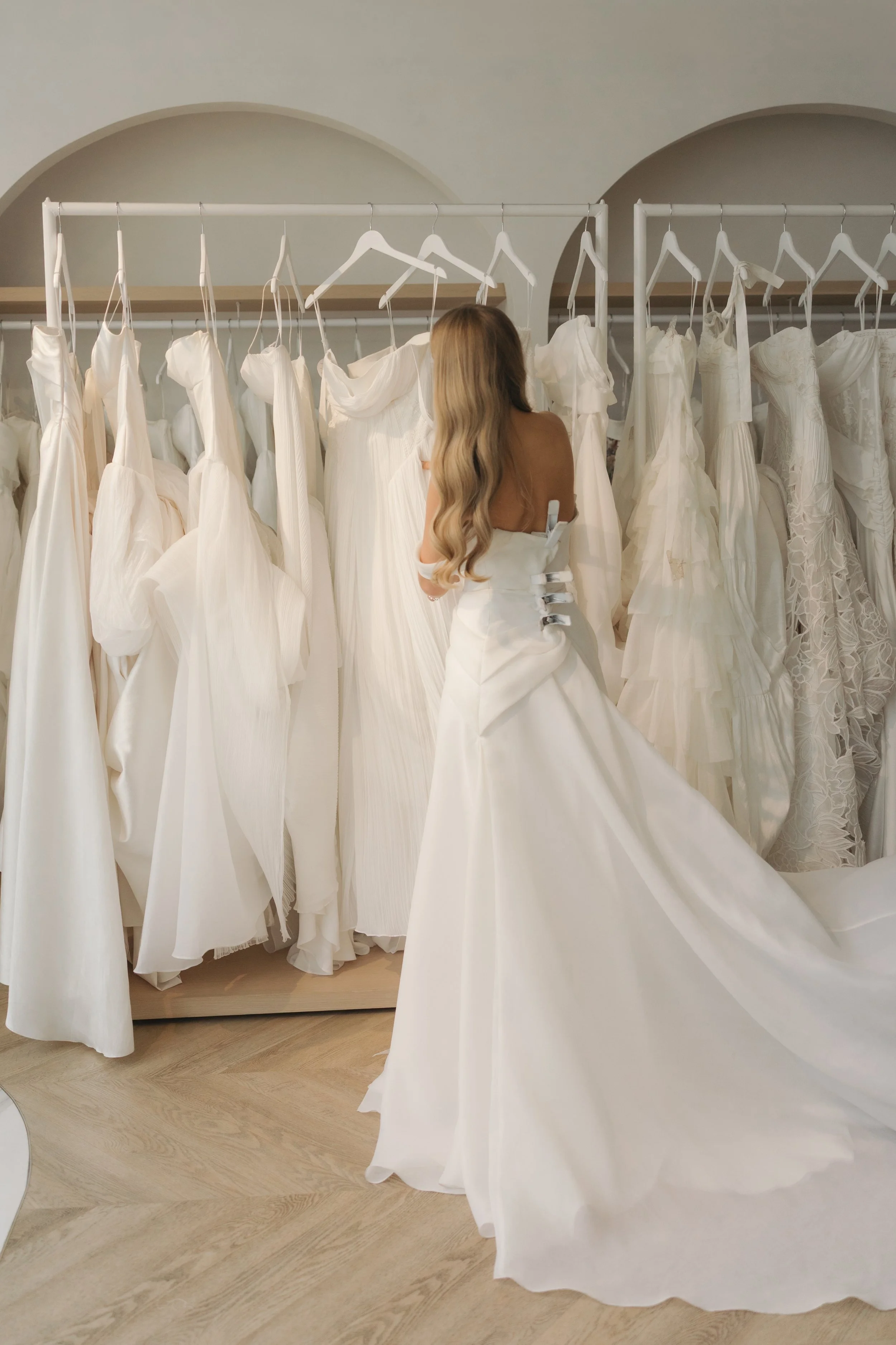 A woman trying on a wedding dress in a bridal boutique, surrounded by racks of white wedding gowns.