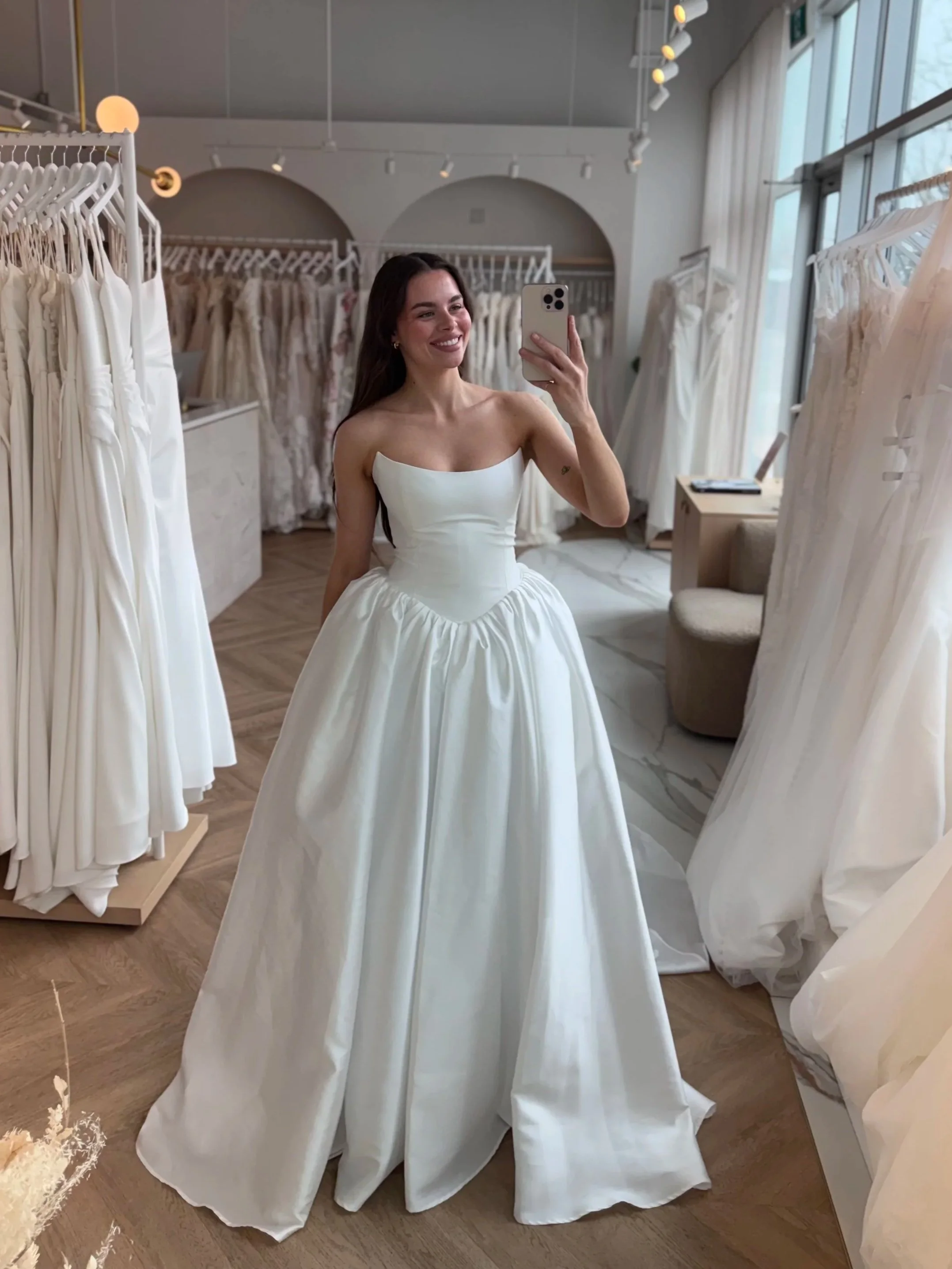 A beautiful bride taking a selfie at The Bridal Boutique in a clean sharp mikado ball gown with a scoop neckline and basque waist. SO stunning. At the bridal boutique with stunning dresses hanging in the background
