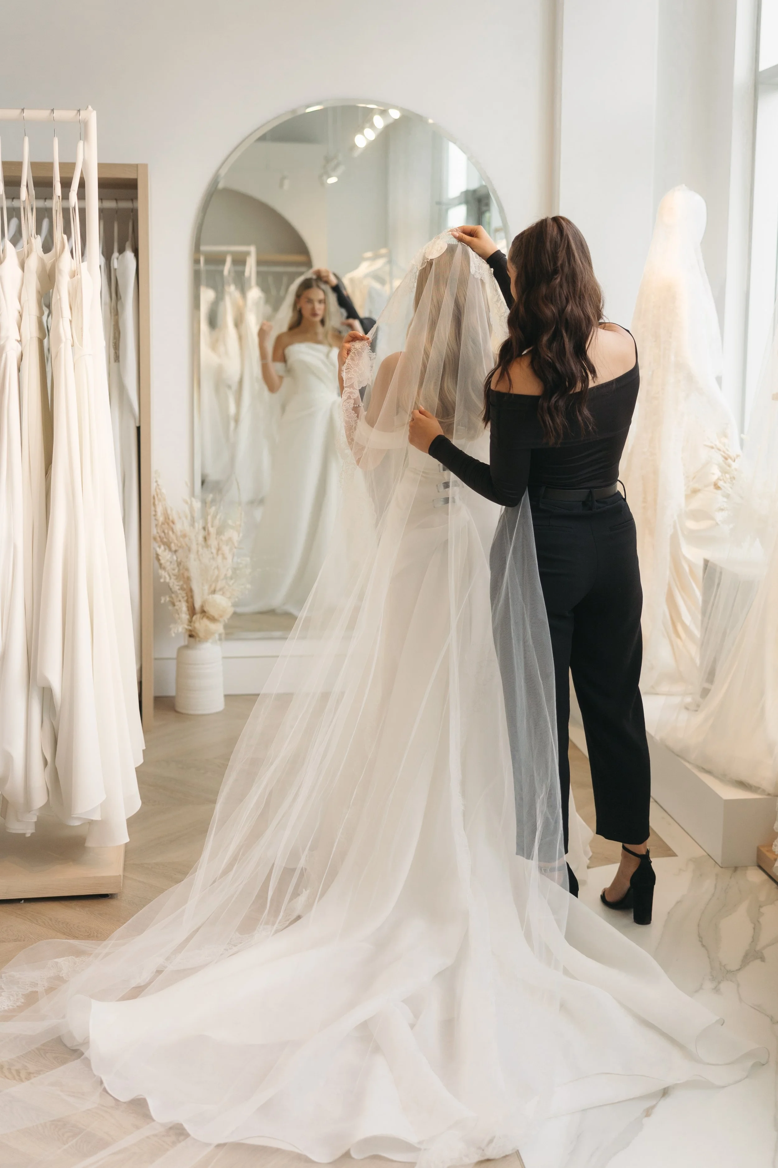 A woman helping a bride try on a wedding dress in a bridal boutique, with a mirror reflecting the bride's face and her wedding gown.