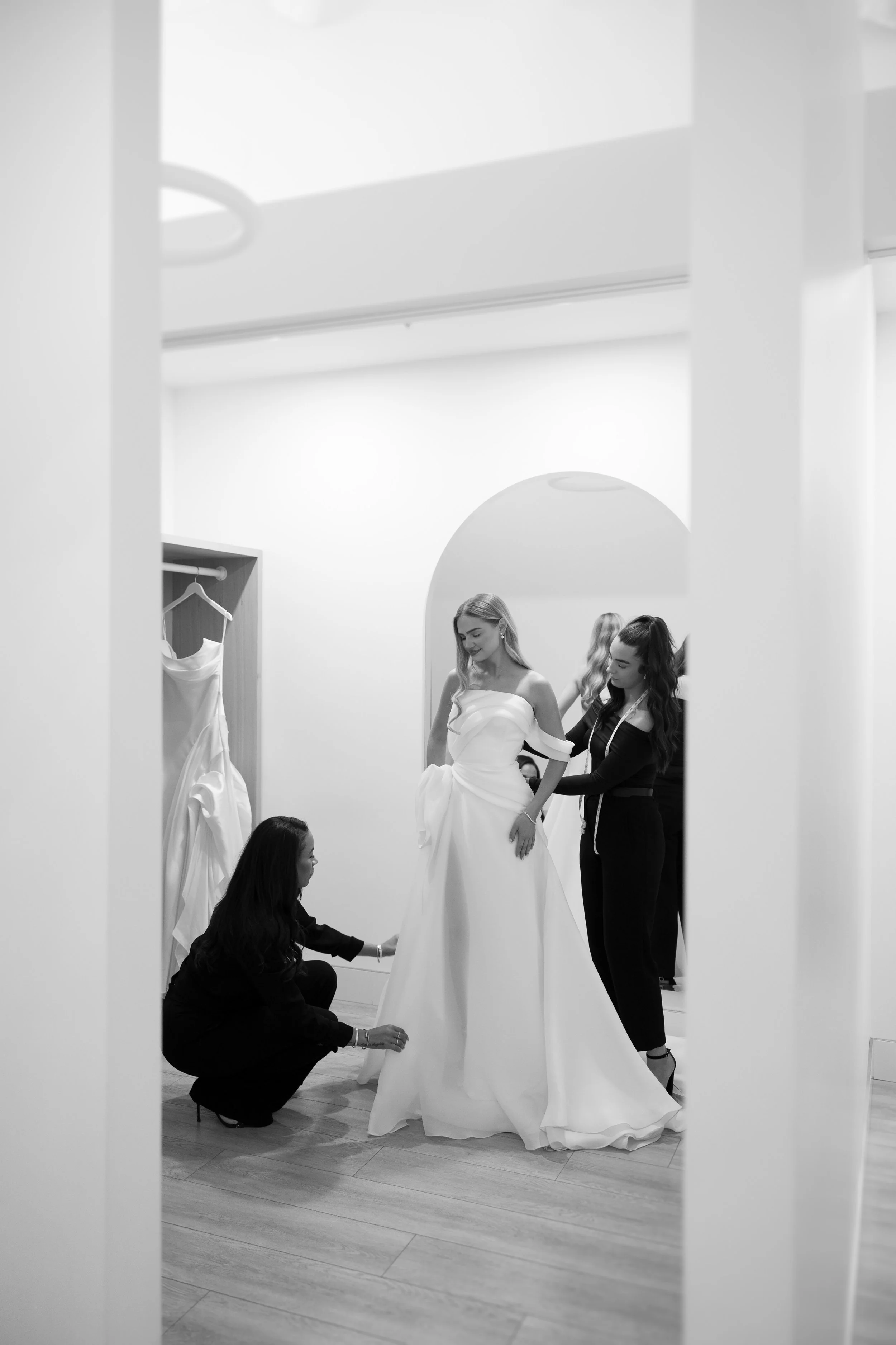 Women helping bride try on wedding dress in fitting room.