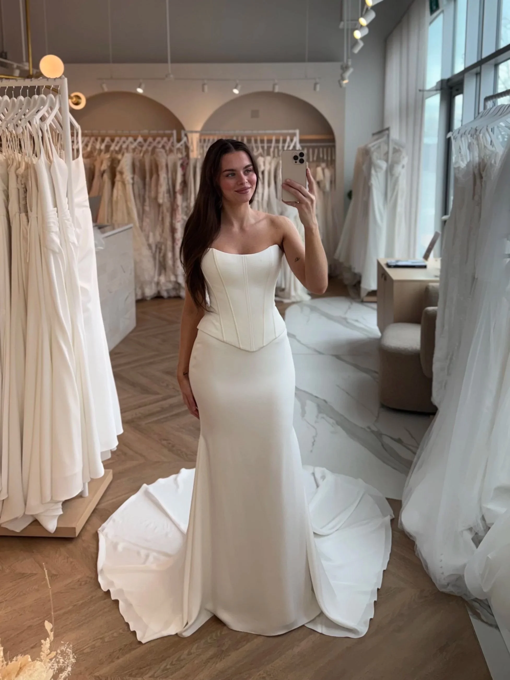 A beautiful bride taking a selfie in a large arched mirror at The Bridal Boutique. She is wearing Paloma Blanca P5160 which is a charmeuse fitted faux 2-piece Basque waist gown with clean lines and super snatched. Stunning dresses hanging in the back