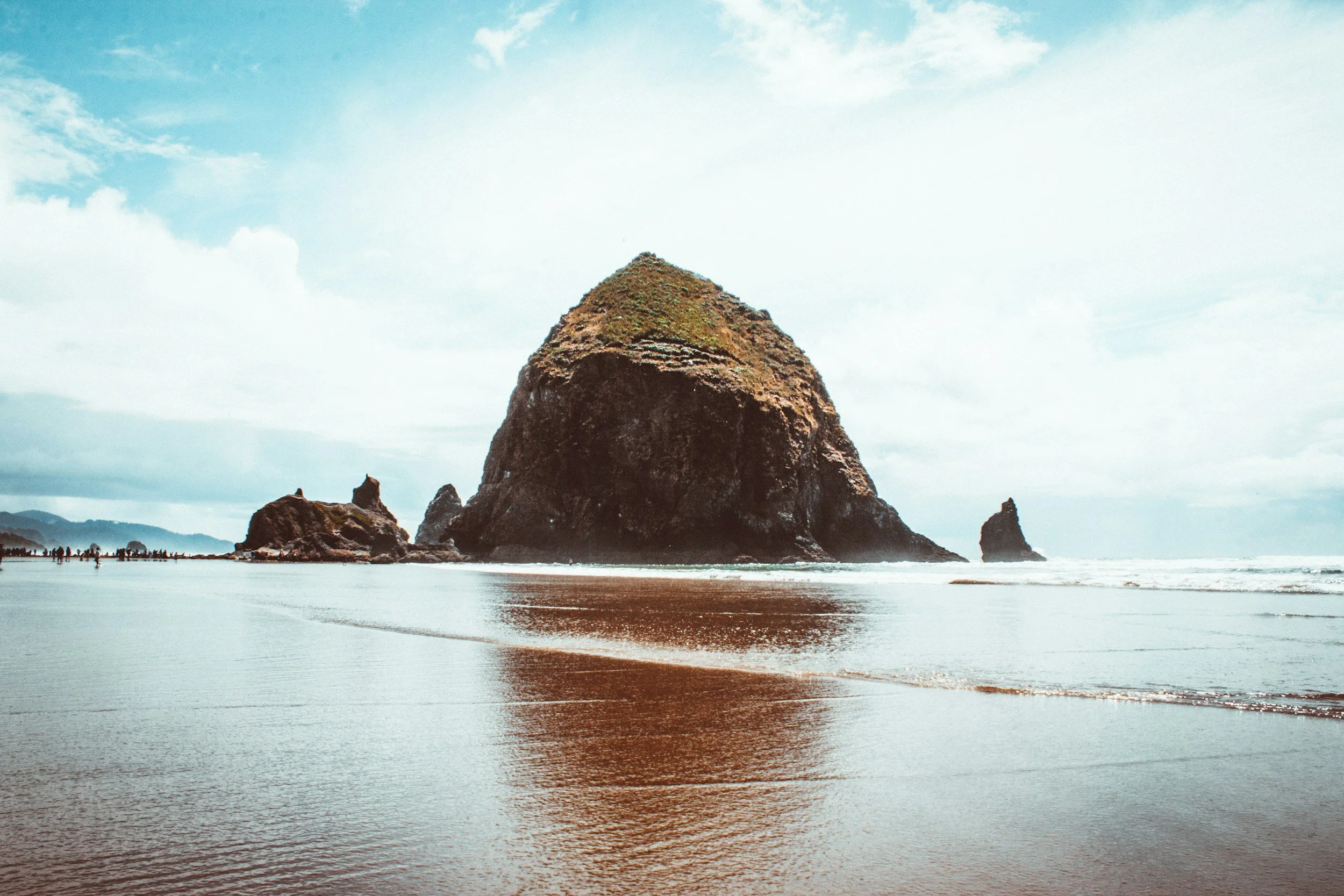 A large rock formation rises from the ocean near the shoreline with smaller rocks around it, and a line of people walking along the beach in the distance under a partly cloudy sky.