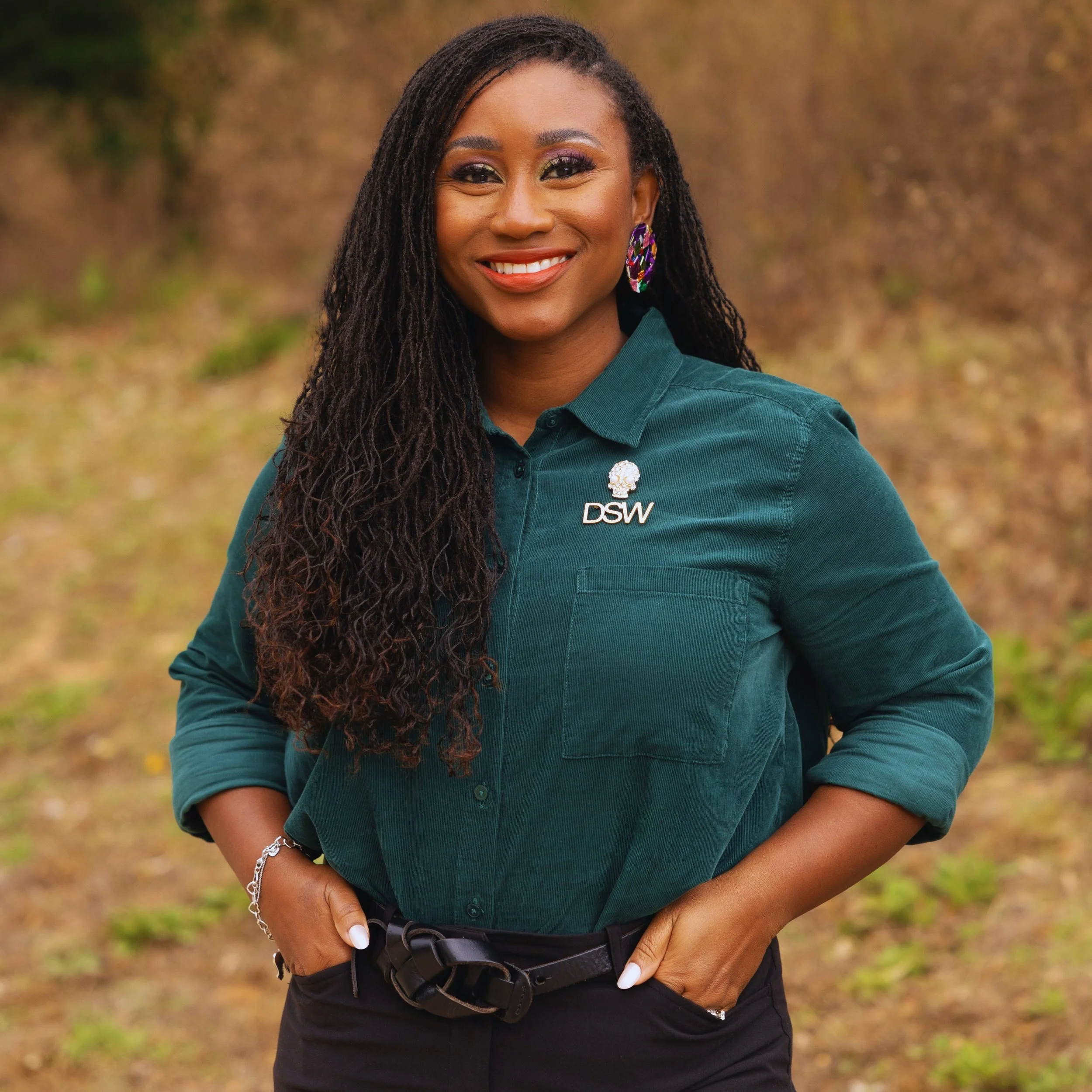 A woman with long, dark, textured hair, smiling outdoors. She is wearing a green button-up shirt with roll-up sleeves, a silver bracelet, large colorful earrings, and a necklace with a skull charm that has the letters 'DSW'. Her hands are in her pockets, and the background is blurred with earthy tones of autumn foliage.
