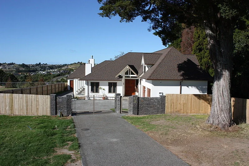 Painted by Europlast Systems in Tauranga, Bay of Plenty. A modern white house with a brown roof, surrounded by a wooden and stone fence, with a gated driveway leading to the house. There is a large tree on the right and a clear blue sky overhead.