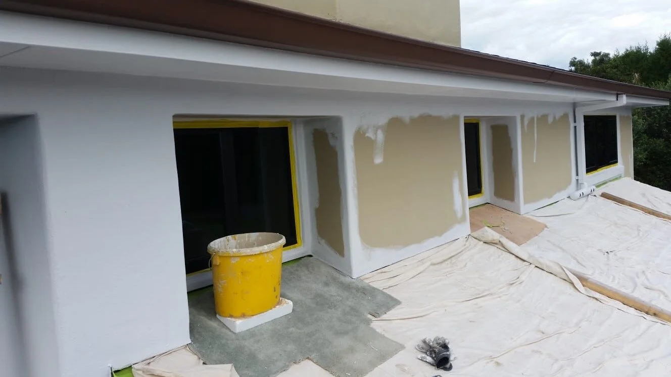 Painted by Europlast Systems in Tauranga, Bay of Plenty. Exterior of a house under renovation with partially painted walls, window frames taped, and construction materials on the ground.