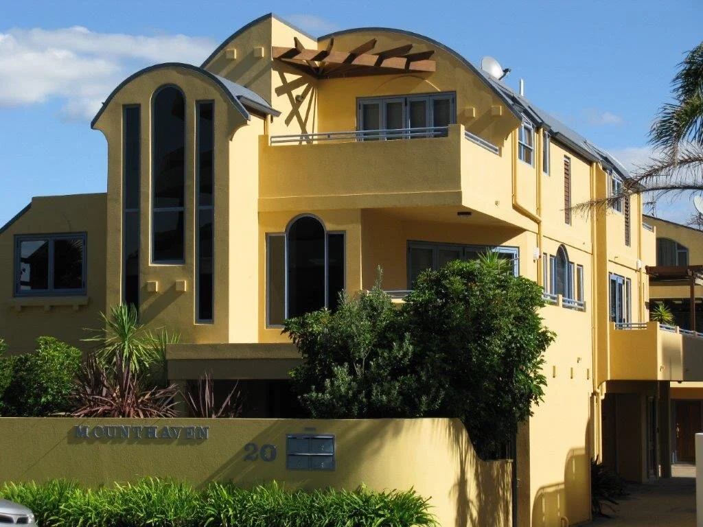 Yellow Tauranga multi-story house with modern architecture, teal window frames, and a sign reading 'Montclaven' surrounded by lush green plants and palm trees painted by Europlast Systems in Tauranga, Bay of Plenty.