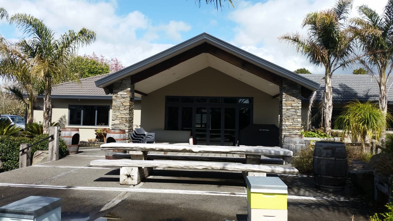 Painted by Europlast Systems in Tauranga, Bay of Plenty. A building with a sloped roof and stone pillars on a cloudy day. There are palm trees and shrubs in front, with wooden benches and barrels around a patio area.