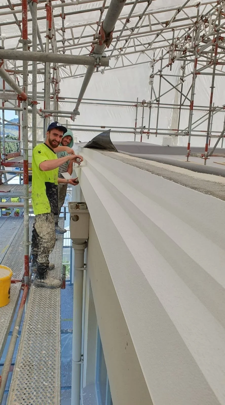 Two construction workers standing on scaffolding working on the exterior of a building. One is wearing a neon yellow safety shirt and the other a hoodie. They are working on a large concrete ledge.