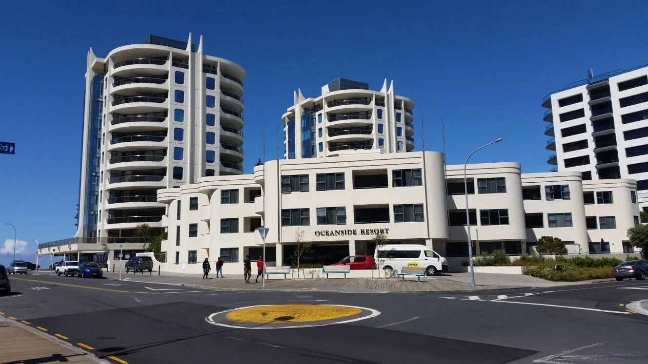 Painted by Europlast Systems in Tauranga, Bay of Plenty. Modern white multi-story hotel building with rounded balconies, labeled 'Oceanside Resort,' situated along a street with cars, pedestrians, and a blue sky.