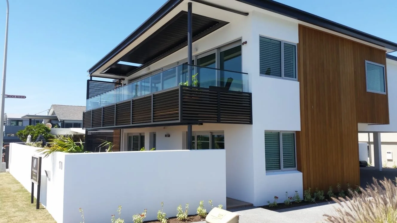Tauranga painting and plastering project of Europlast Systems of a modern two-story house with white walls, wooden accents, and large windows. The house has balconies with glass railings and a small garden area in front.