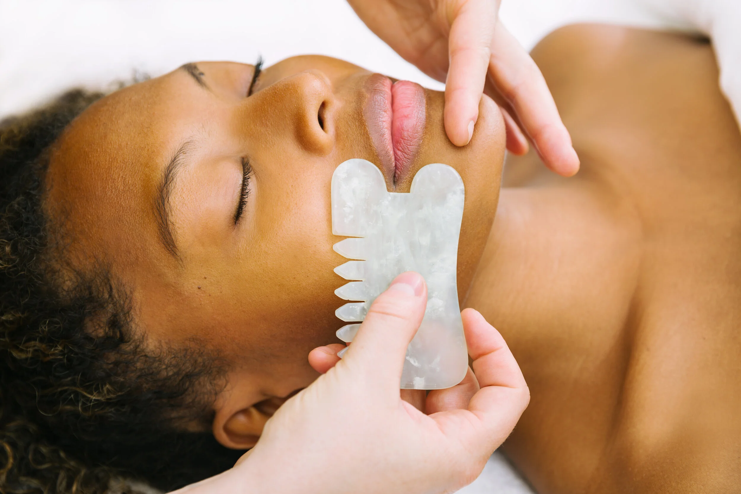 A woman receiving a jawline massage with a gua sha stone on her face while lying down with her eyes closed.