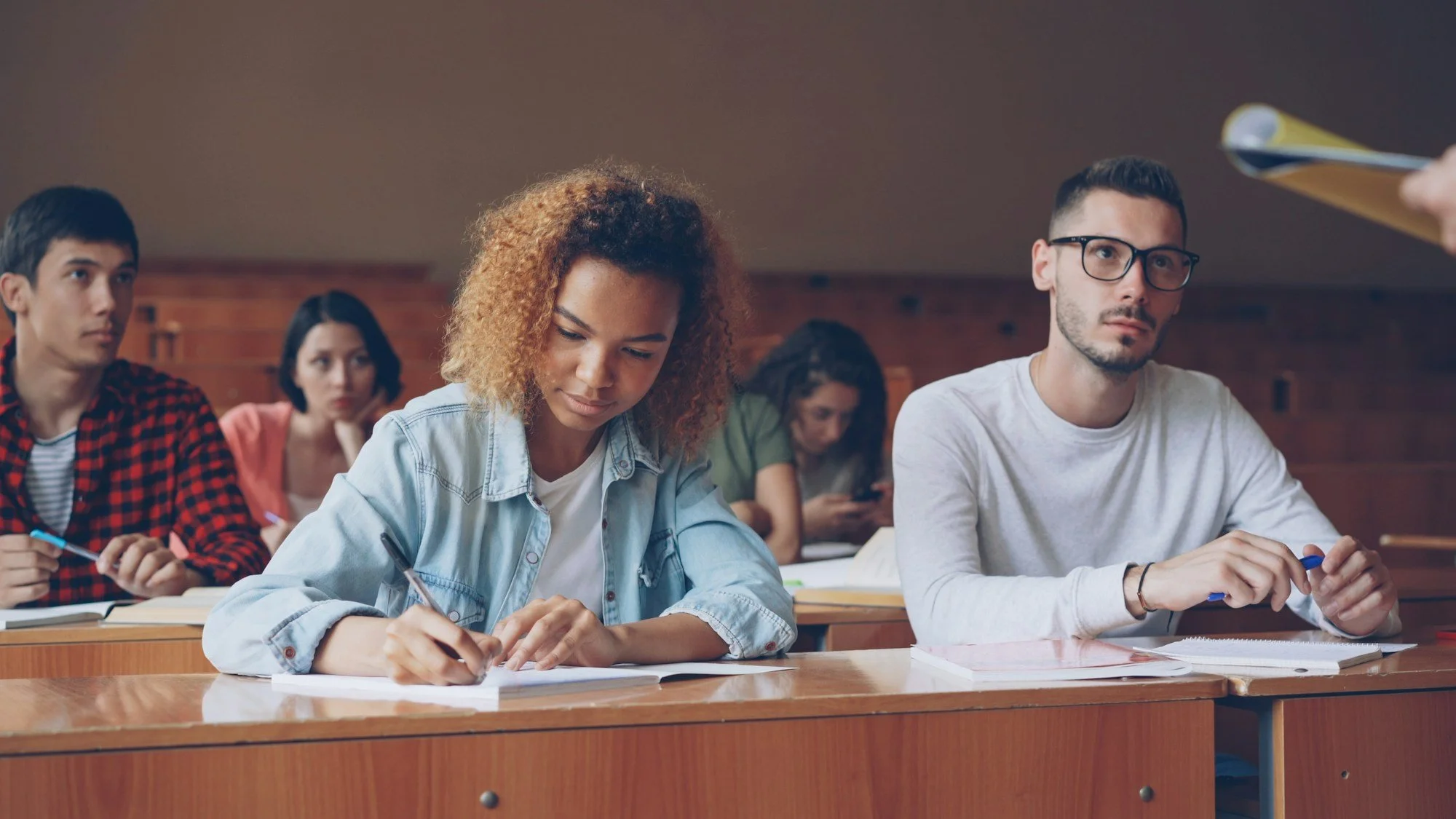 College students seated in rows in a lecture hall, listening and taking notes during a class.