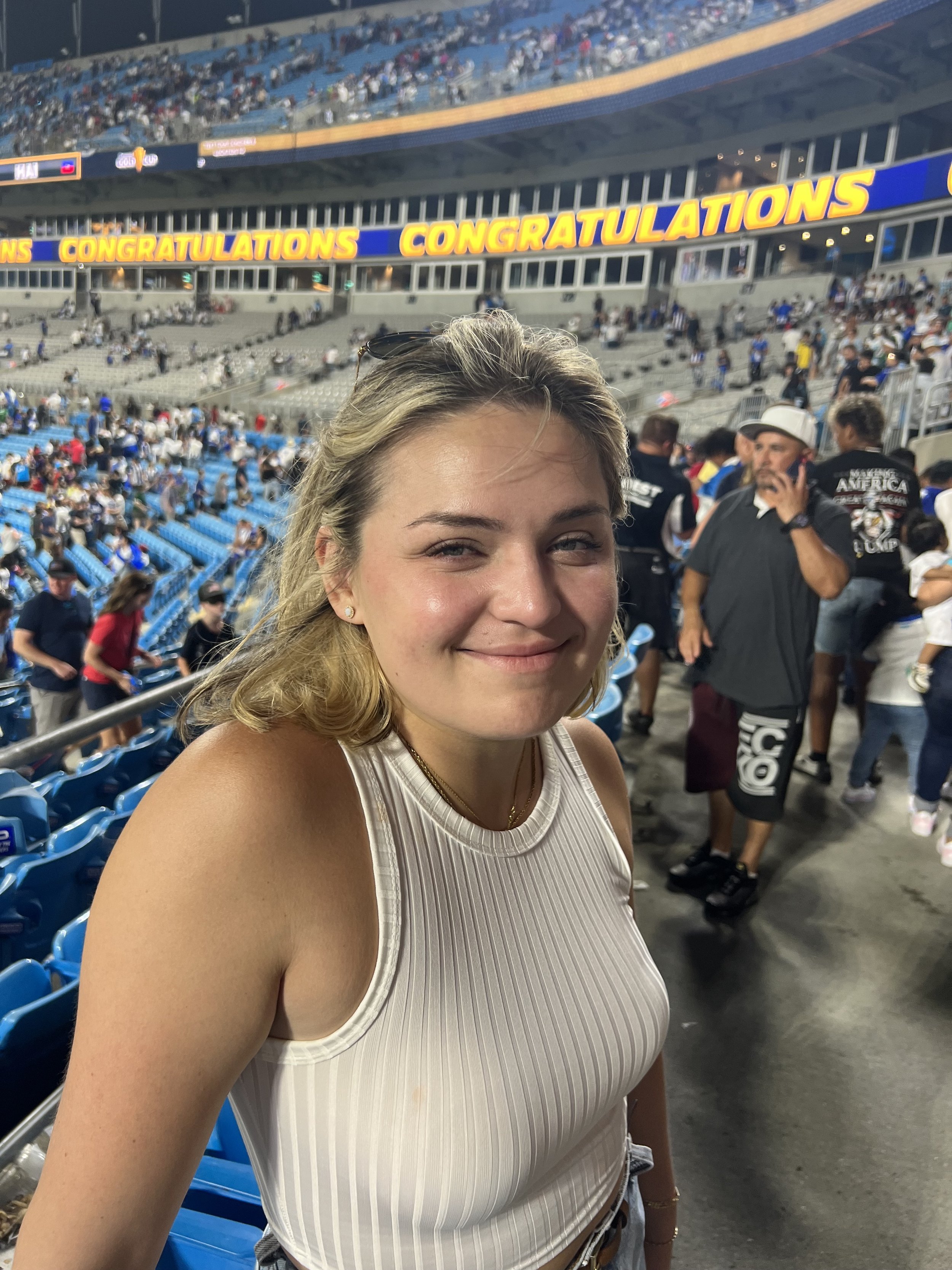 A woman sitting on a sofa smiling and pointing at a birthday cake with a lit sparkler, surrounded by flowers and drinks.