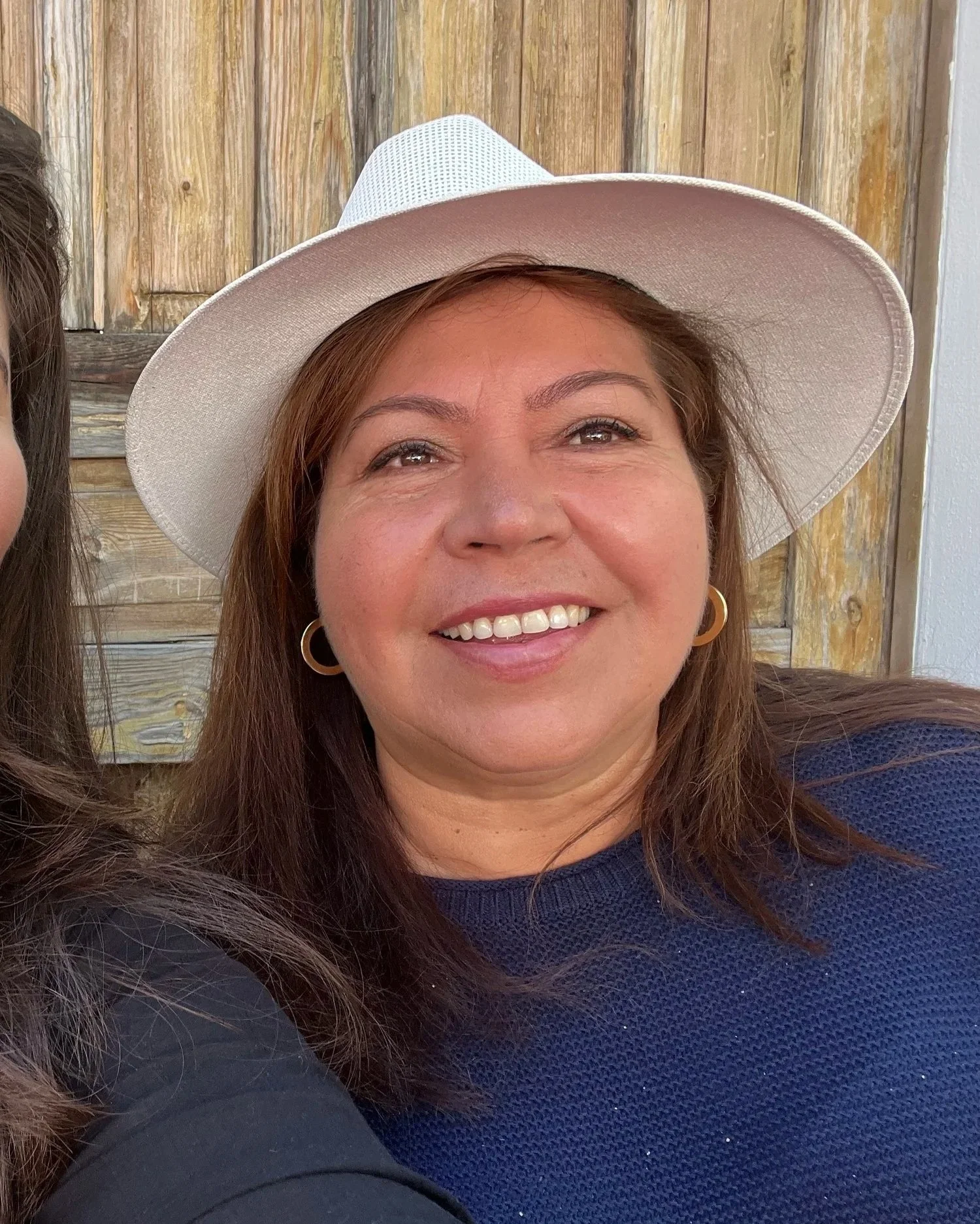 Smiling woman with dark hair, wearing a blue and white striped shirt with tied sleeves, holding a large floppy sun hat in a retail store aisle marked 'beauty' and 'intimates.'