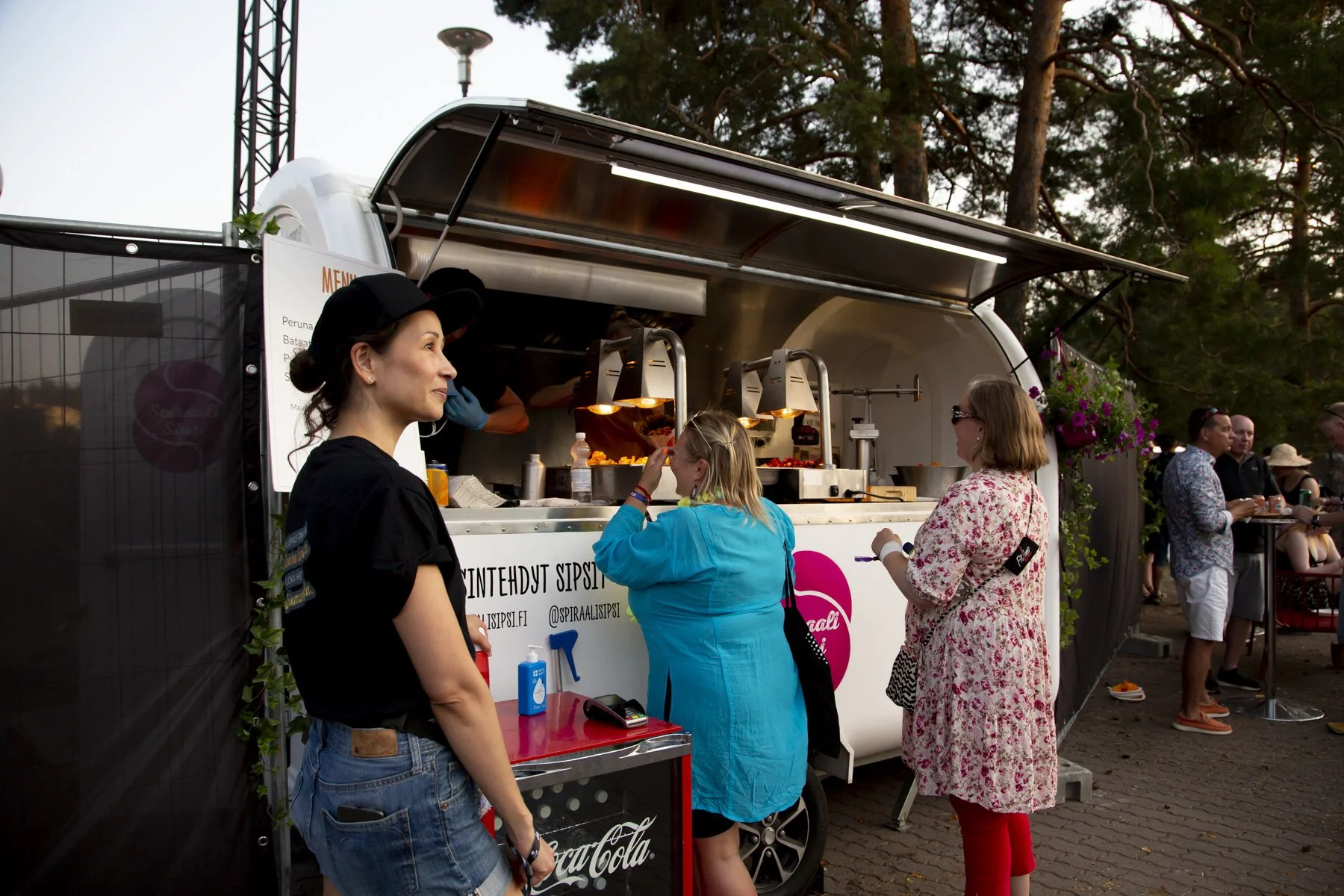 Ruokarekka puistossa, ihmisiä ostamassa ruokaa, pöydät kukkien kanssa ympärillä.