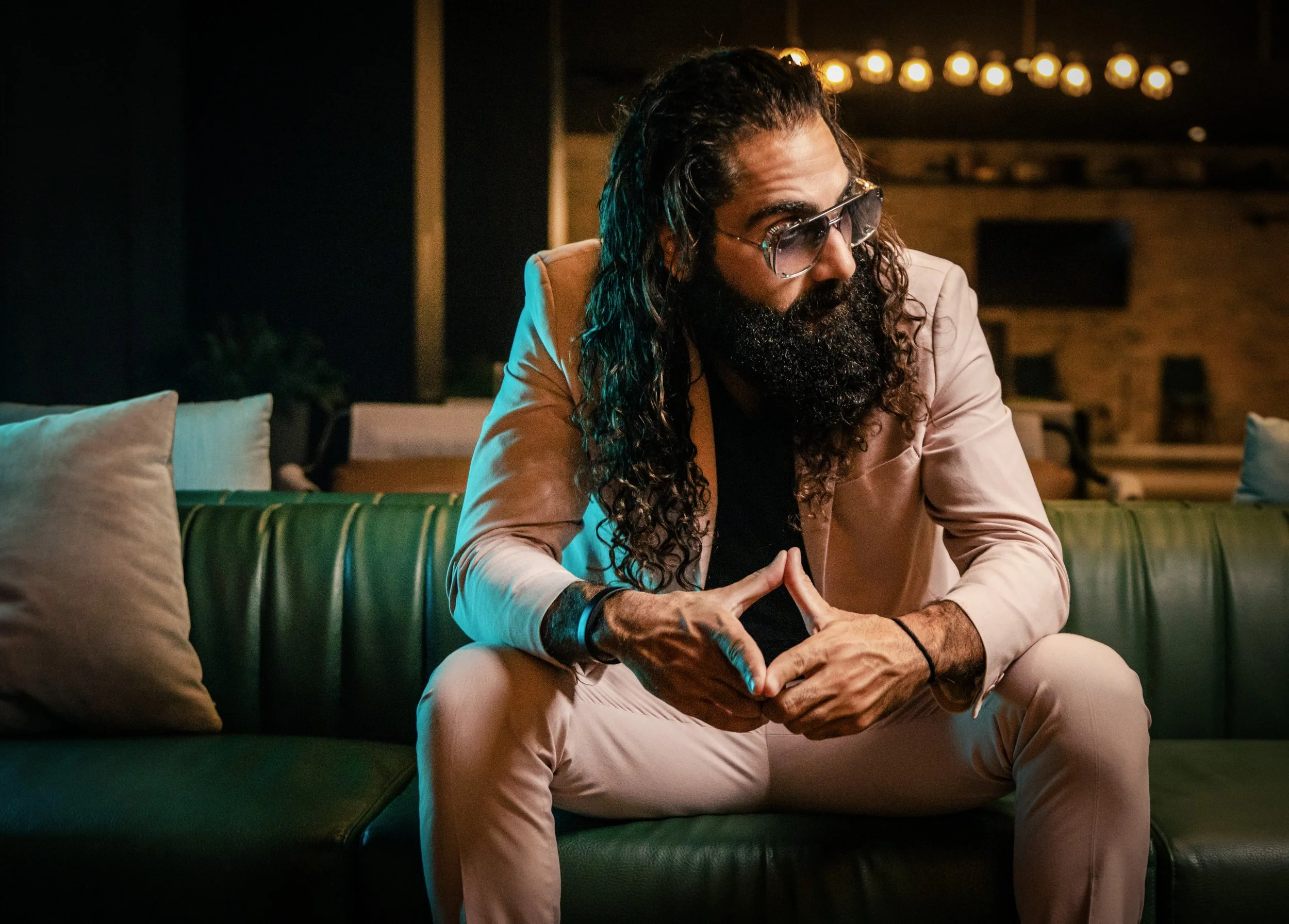 A man with long hair, a beard, sunglasses, and a beige suit is sitting on a green couch in a dimly lit room, with string lights in the background.