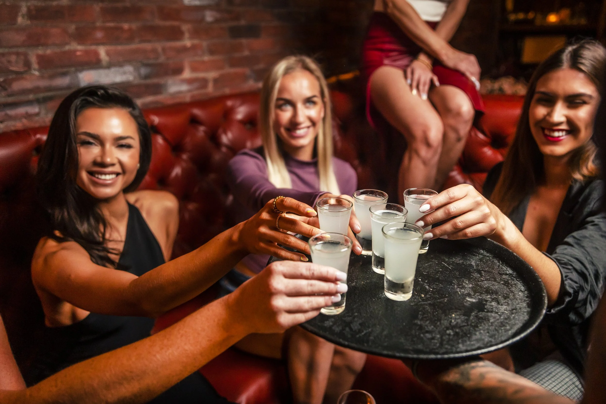 A group of four women sitting on a red leather booth in a bar, clinking shot glasses filled with a clear beverage, smiling and enjoying drinks together.