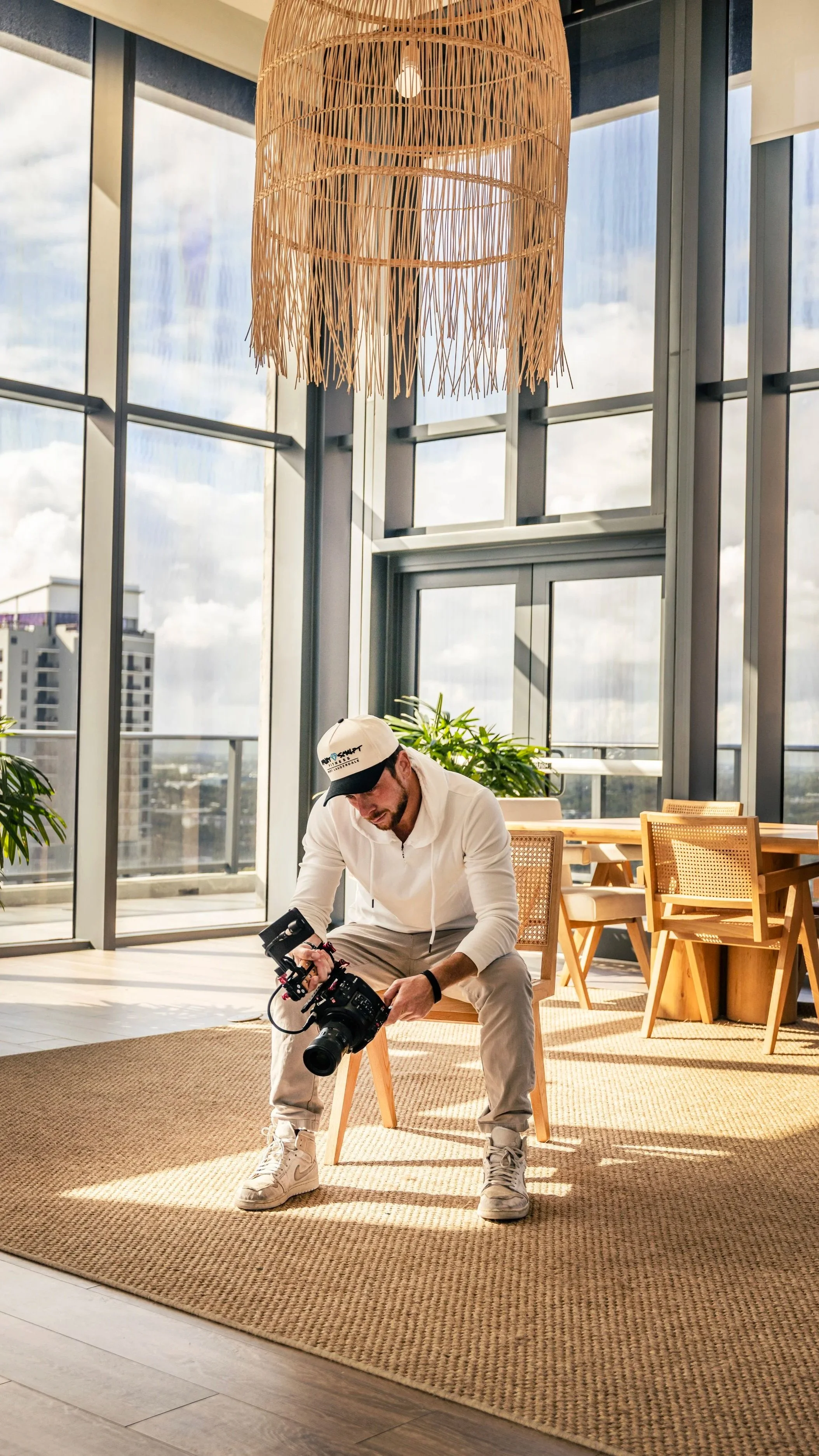 A man in a white hoodie and beige pants is sitting and adjusting a professional camera in a bright, modern room with large floor-to-ceiling windows, wooden furniture, and an overhead woven light fixture.