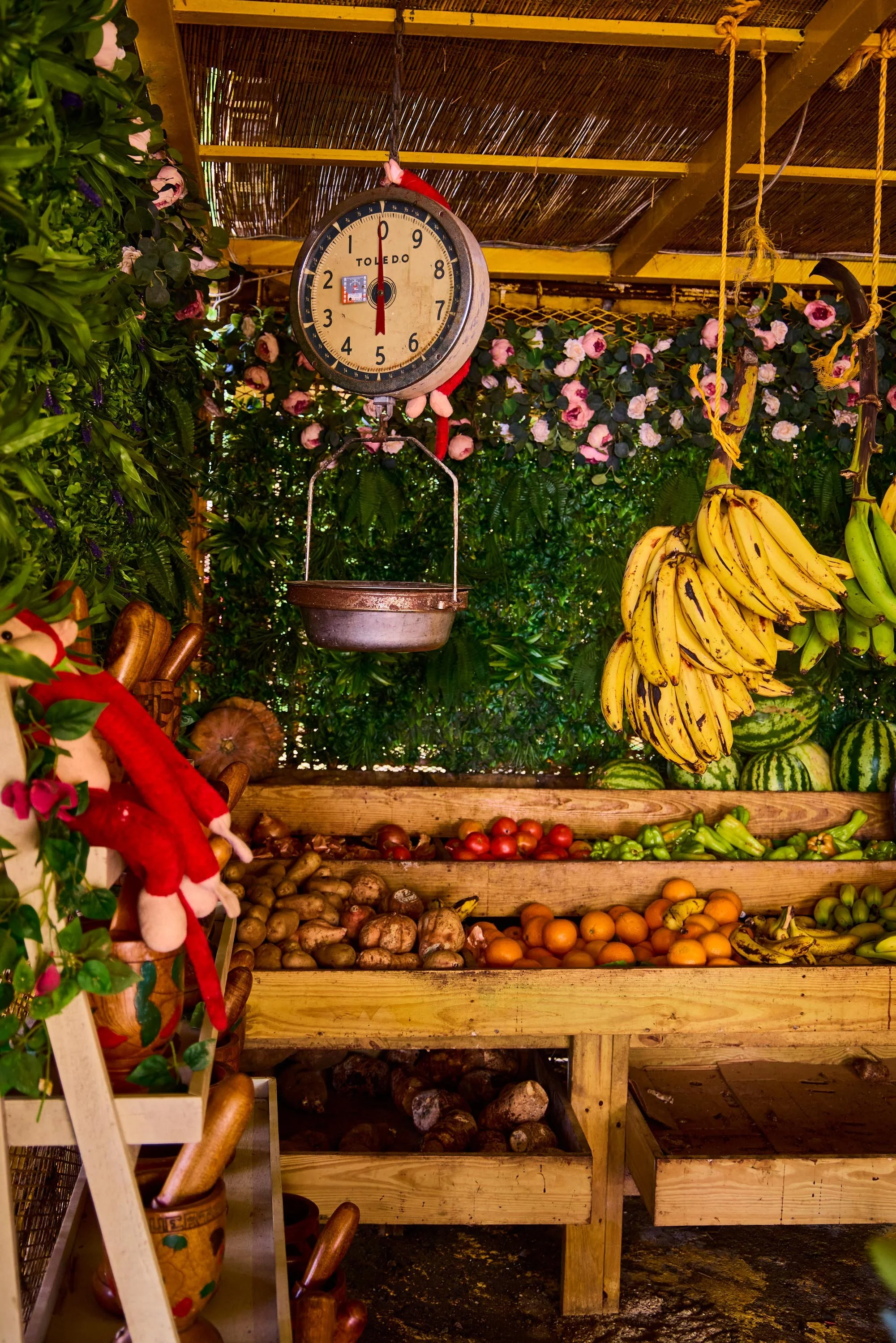 A wooden display with three shelves filled with various fruits and vegetables, including bananas, watermelons, tomatoes, potatoes, and other produce. A vintage clock hangs above the display, surrounded by greenery and pink flowers.