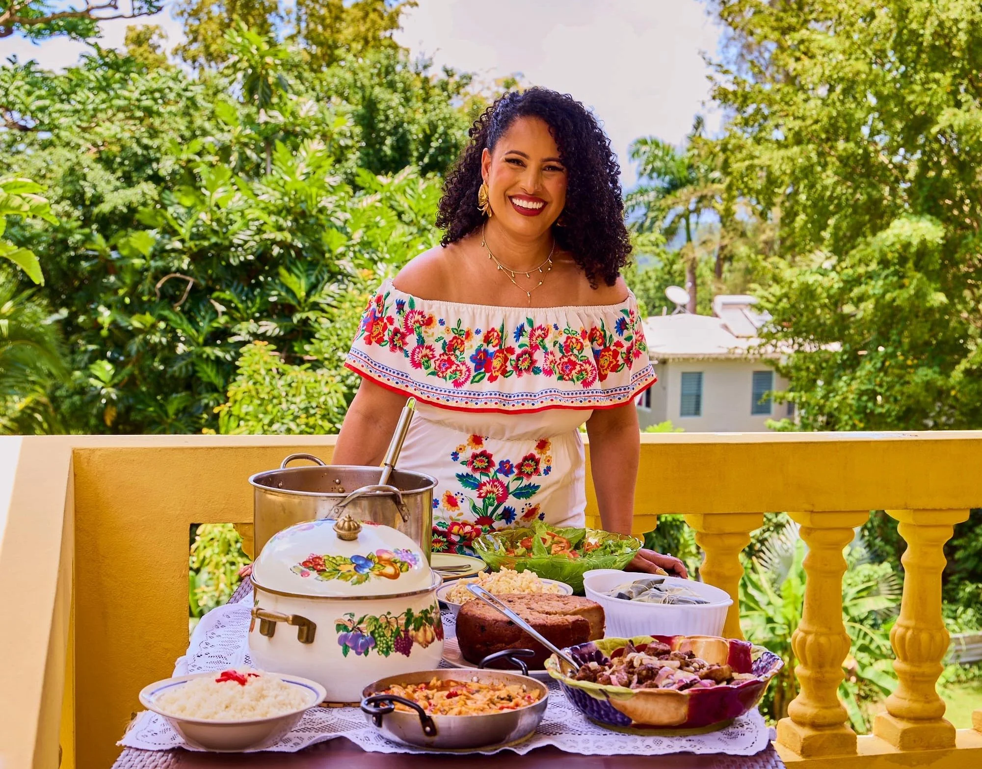 A woman with curly dark hair, wearing a colorful embroidered off-shoulder top, is smiling and standing behind a table of food on a balcony outside. The table has various dishes including salads, a cake, and other cooked foods. There are lush green tr