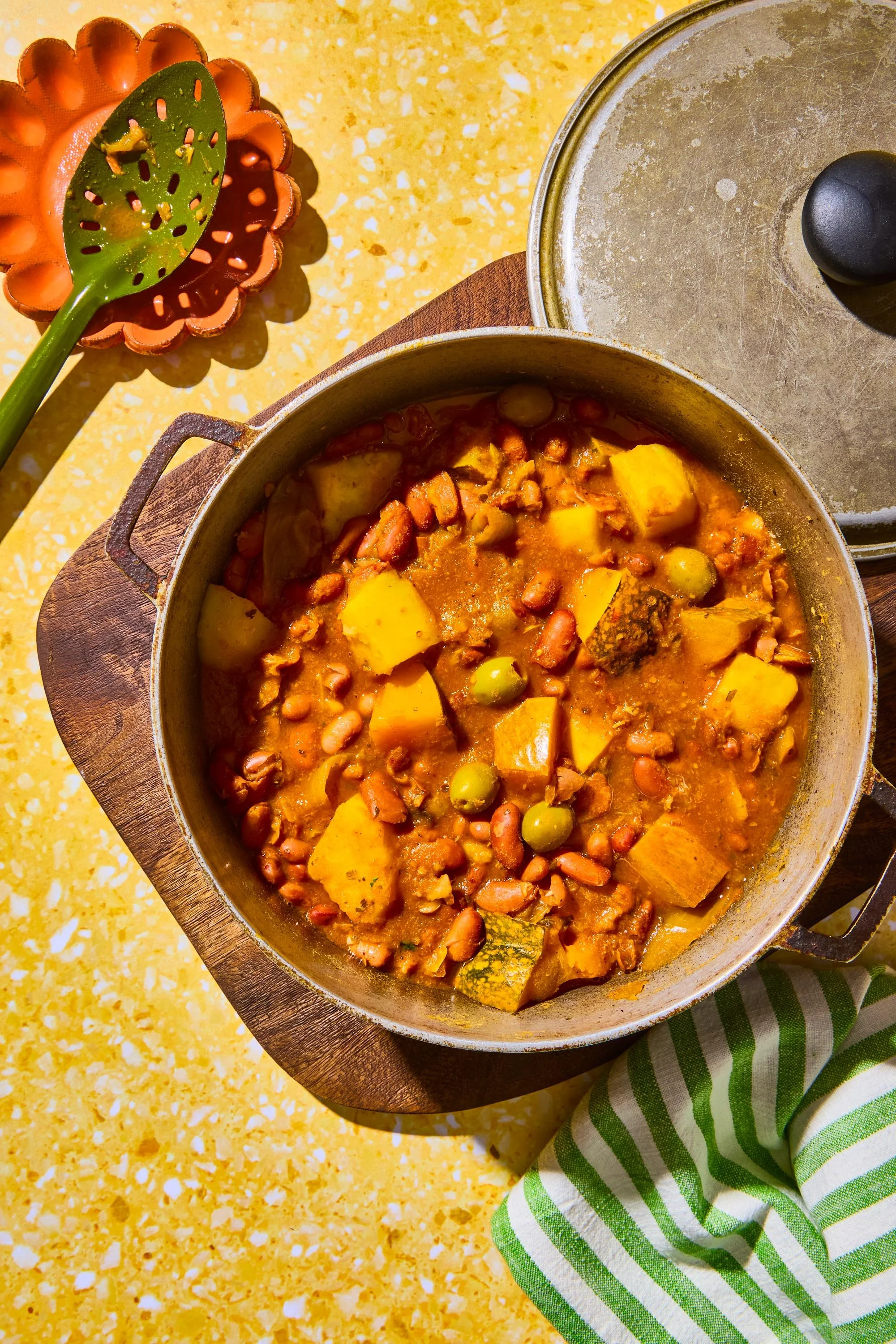 Pot of vegetable and bean stew on a yellow speckled countertop, with a green and white striped cloth nearby, and a decorative orange and green ladle resting on a small orange bowl.