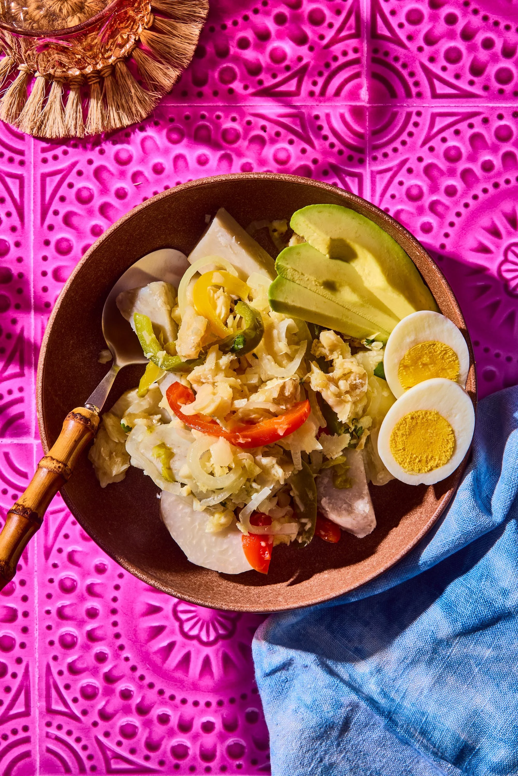 A bowl of potato salad topped with sliced avocado, halved hard-boiled eggs, and vegetable slices, placed on a vibrant pink patterned tablecloth with a blue fabric nearby.