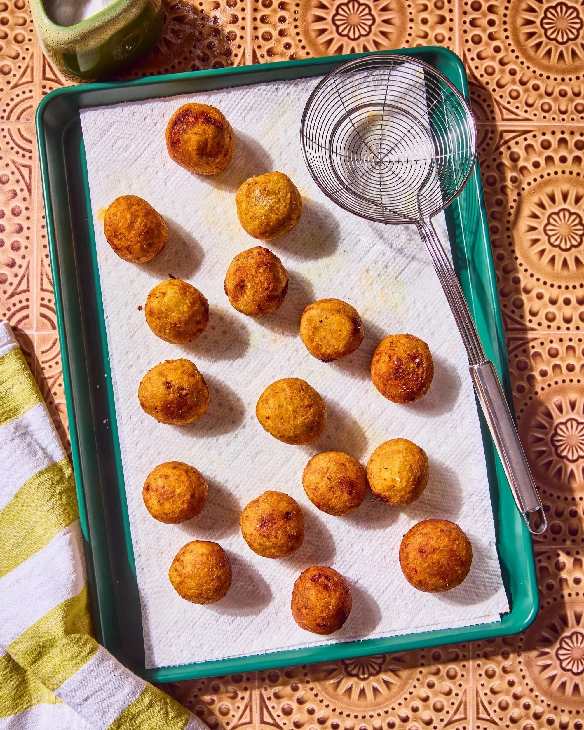 Fried food balls on a paper-lined tray, a wire skimmer resting on the tray, a green drink in a cup, and a yellow and white striped cloth on a patterned brown table.