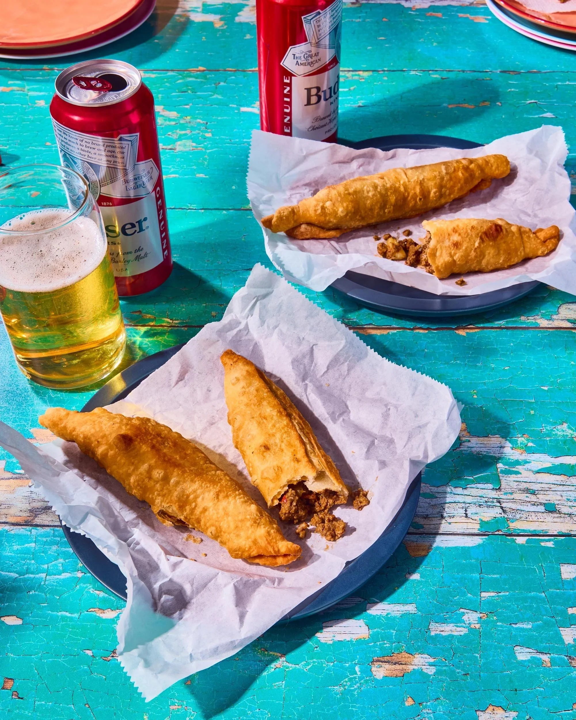 Fried fish fillets on plates, two cans of Budweiser beer, and a glass of beer on a weathered turquoise table.