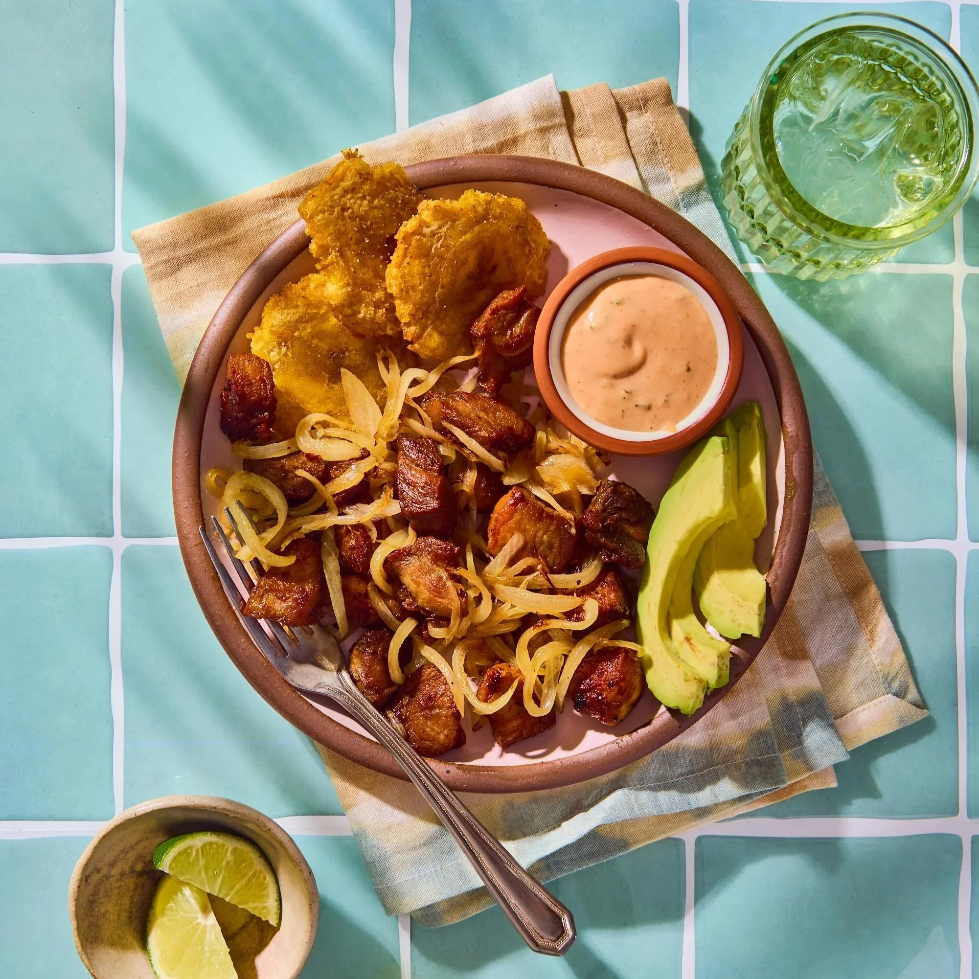 A bowl of Mexican food with fried fish, pork, onions, and a side of avocado slices and a small container of sauce on a light blue tiled table, with lemon wedges on a small dish.