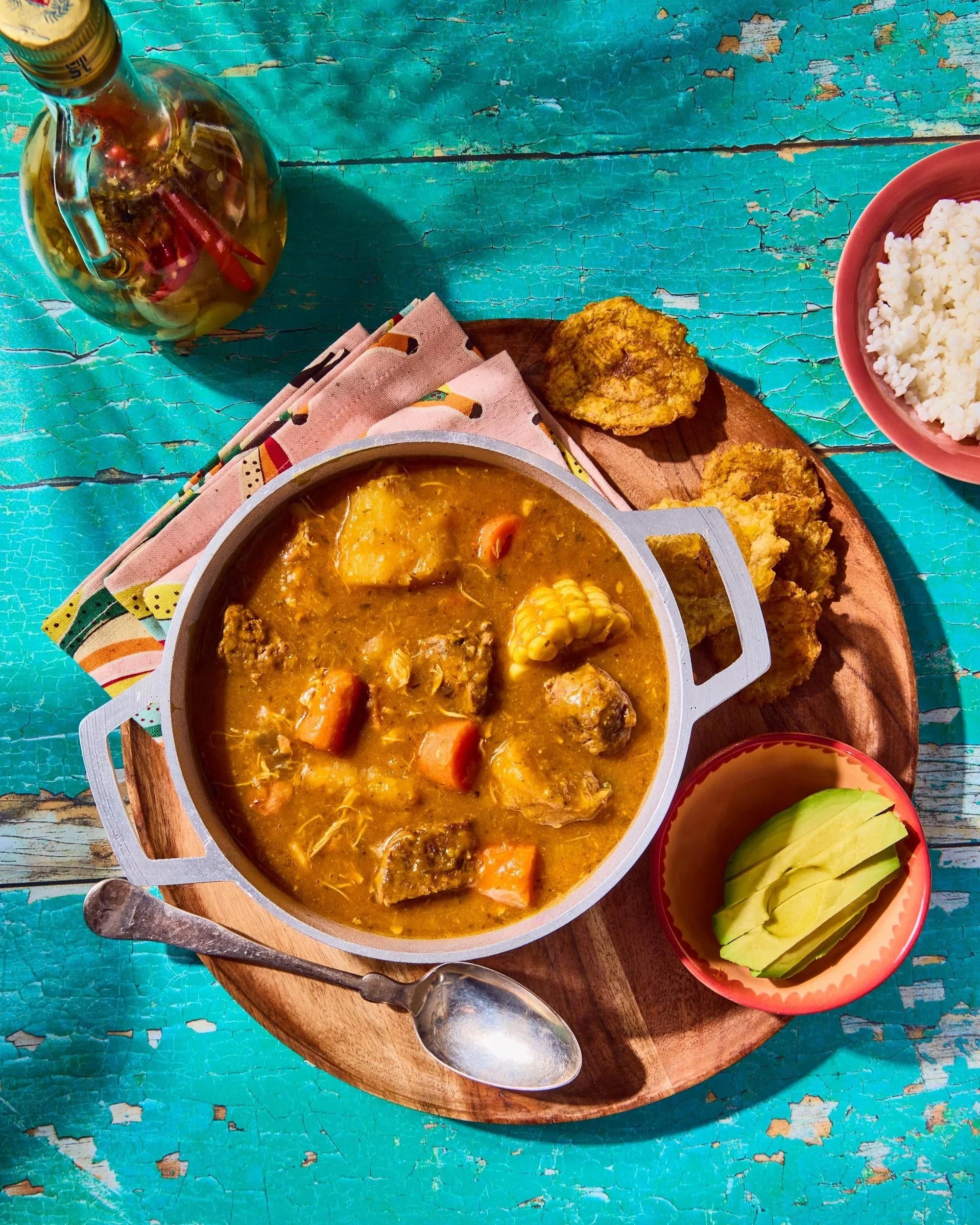 A bowl of hearty stew with vegetables and chunks of meat, accompanied by fried plantains, a small bowl of sliced avocado, white rice in a pink bowl, and a glass bottle of oil on a distressed turquoise wooden table.