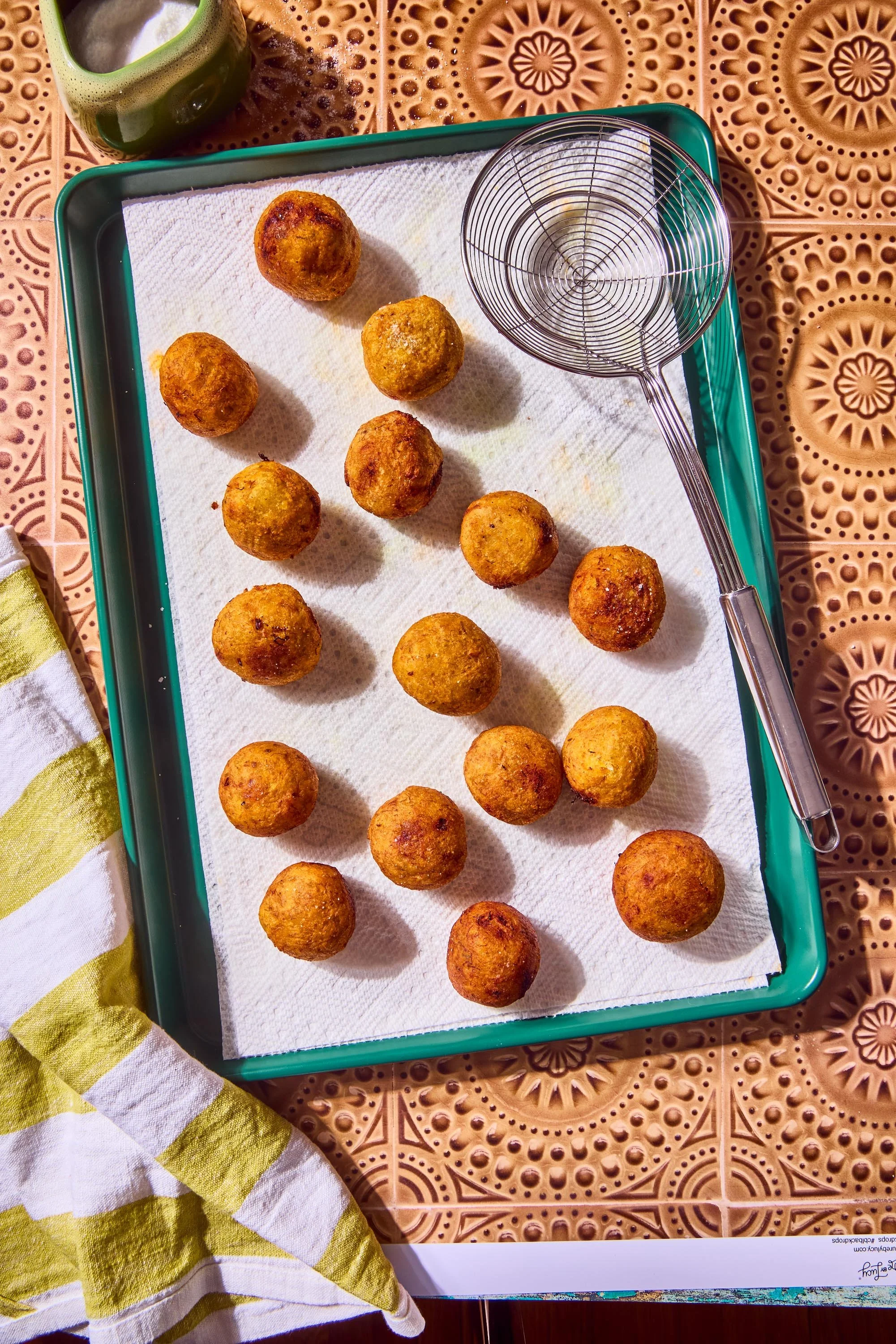 Fried food balls on a paper-lined tray, a wire skimmer resting on the tray, a green drink in a cup, and a yellow and white striped cloth on a patterned brown table.