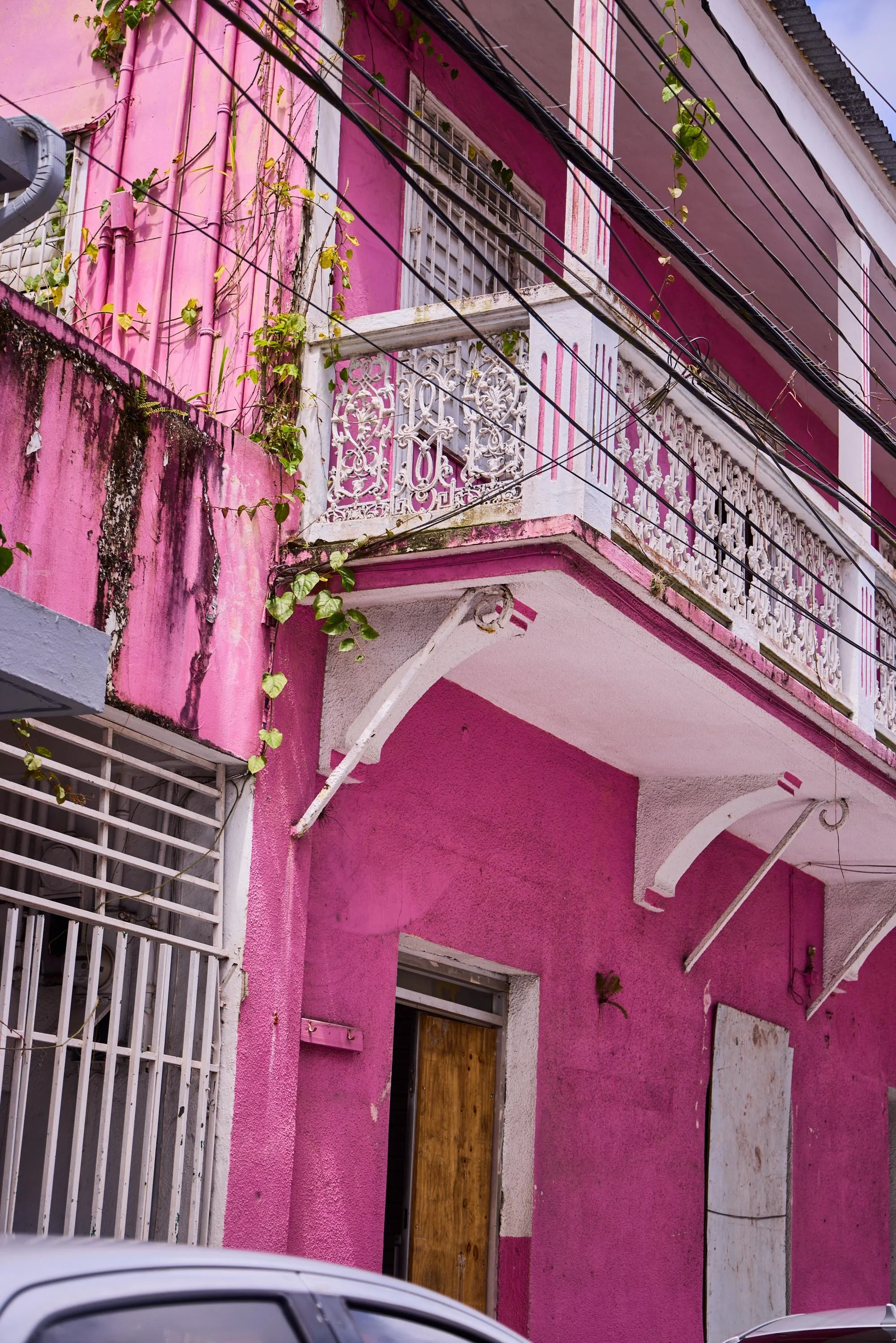 A pink building with ornate white balconies and window grills. The wall shows signs of wear with some dirt and peeling paint. There are black electrical wires crossing the scene. A street with parked cars is visible in the lower part of the image.