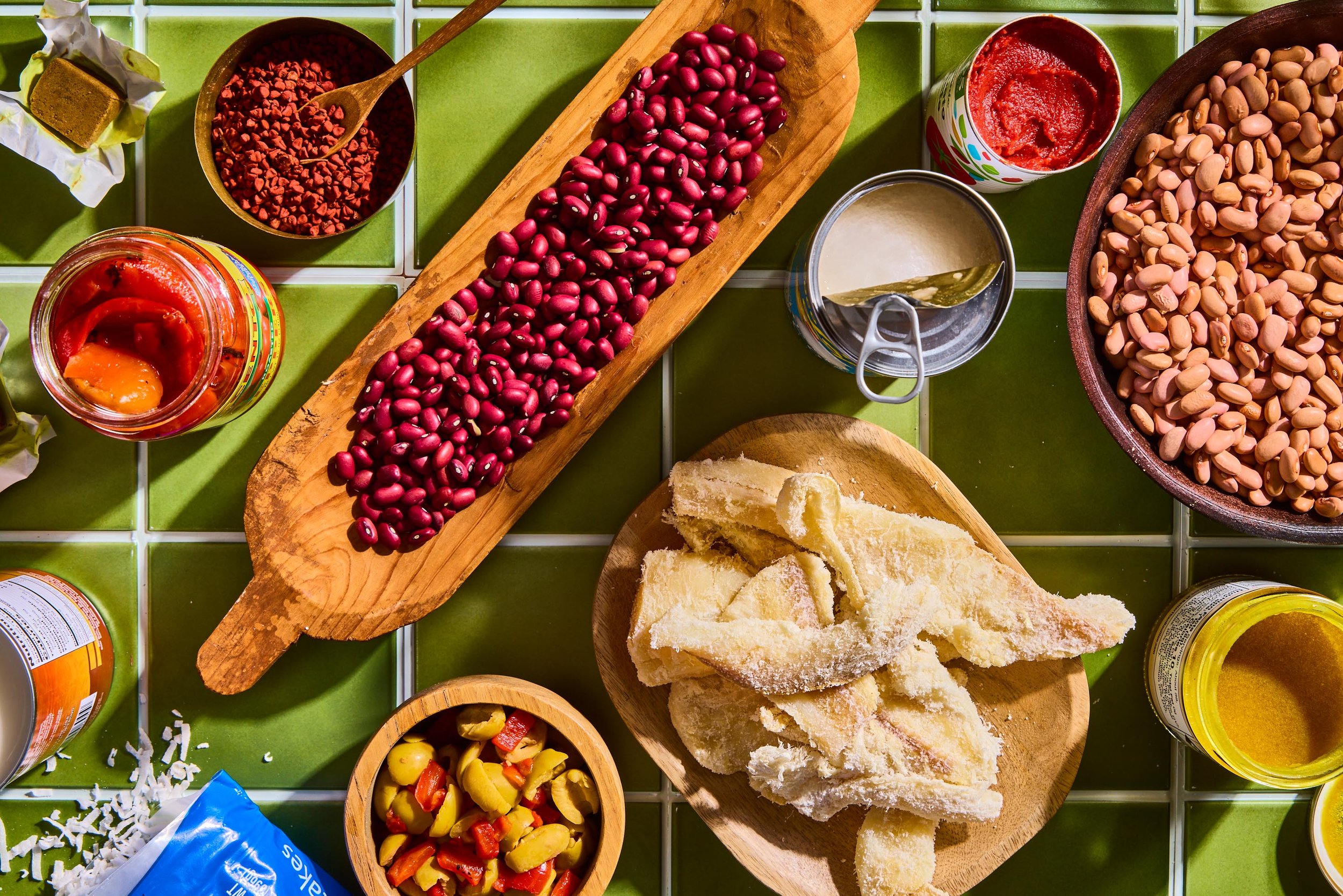 A variety of food items on a green tiled surface, including beans, pasta, canned tomatoes, and bread.