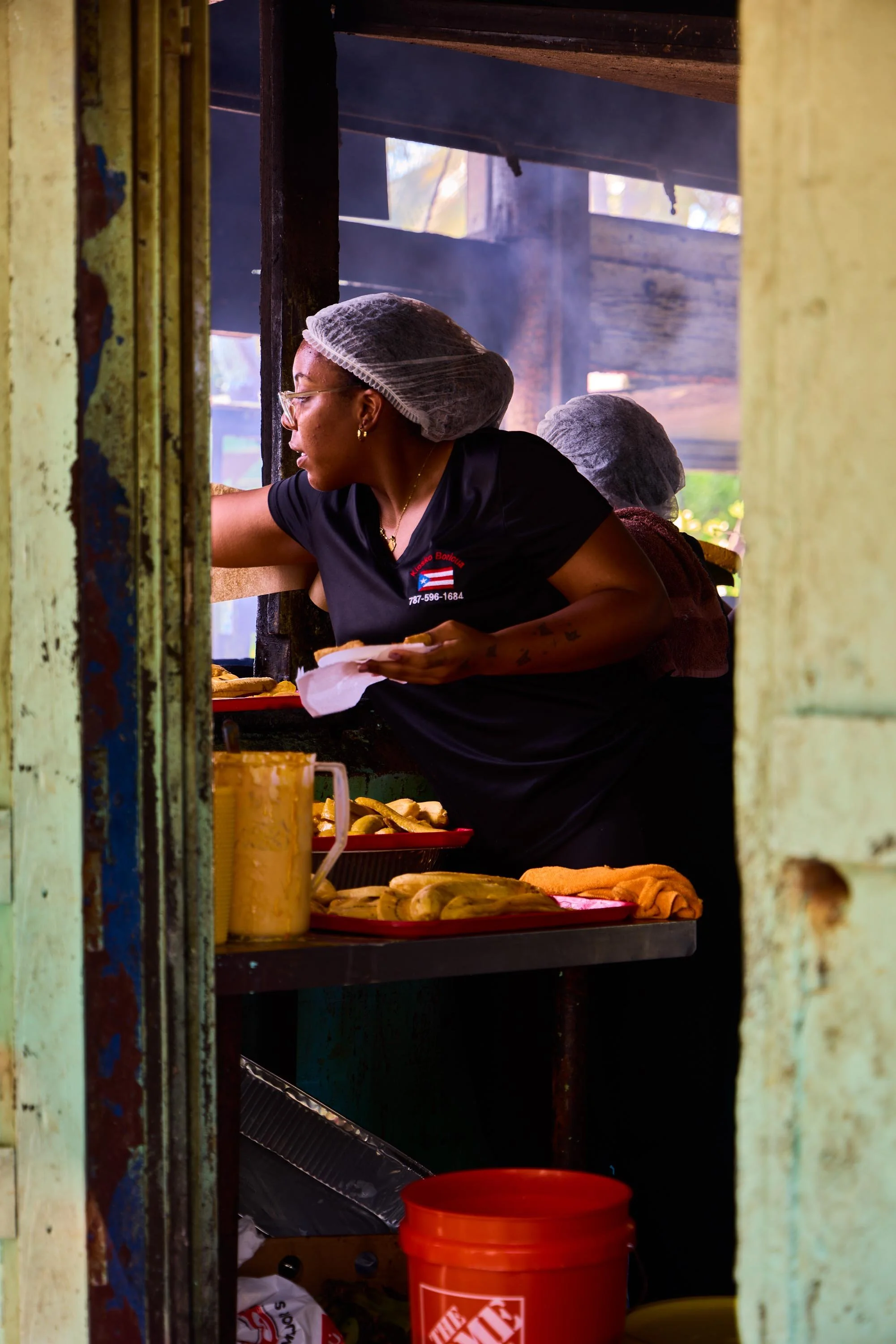 A woman wearing a hairnet, glasses, and a black shirt working at a food stand, serving food, with trays of hot dogs and condiments in front of her.
