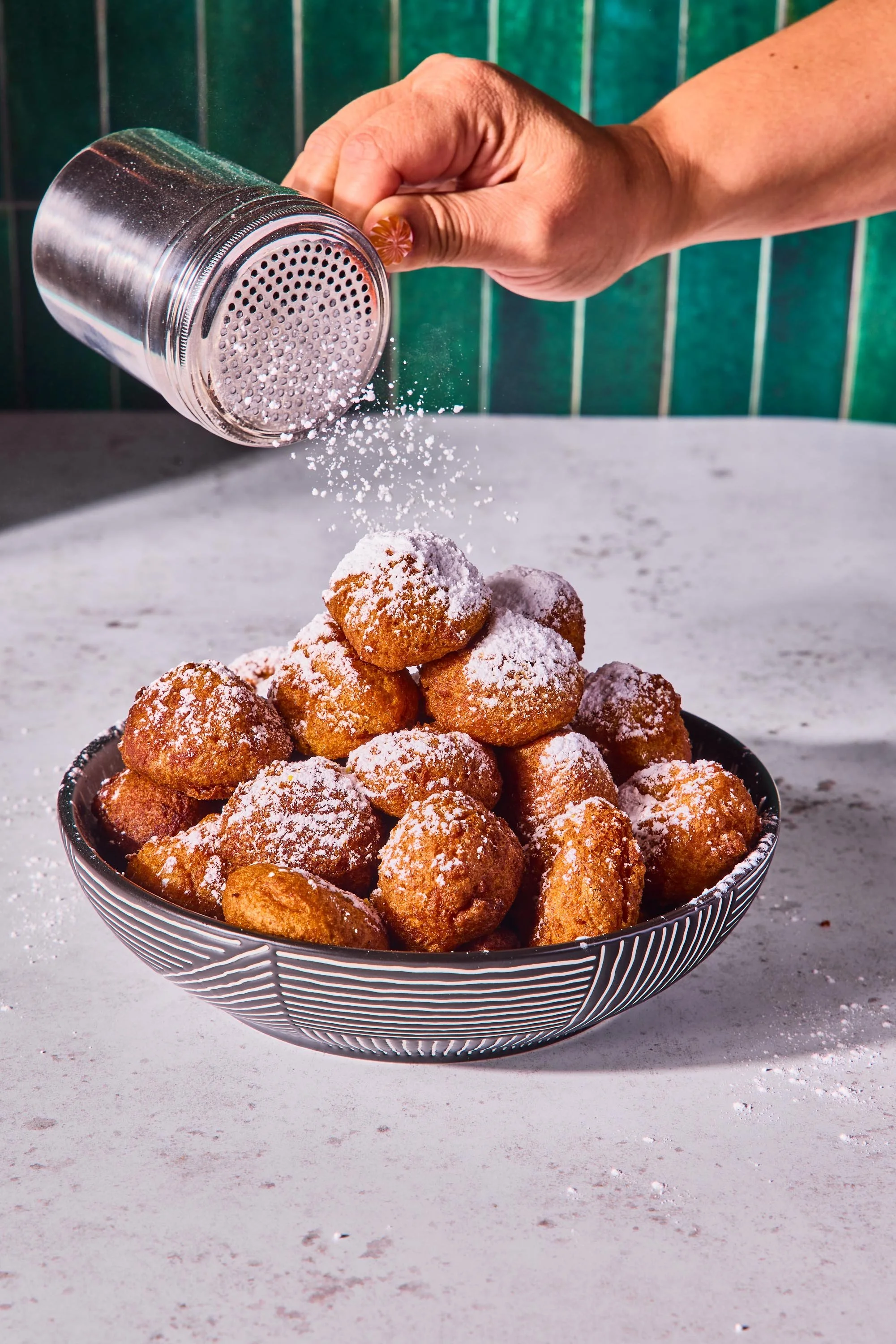 A bowl of fried dough balls dusted with powdered sugar, with powdered sugar being sprinkled over them from a glass shaker.