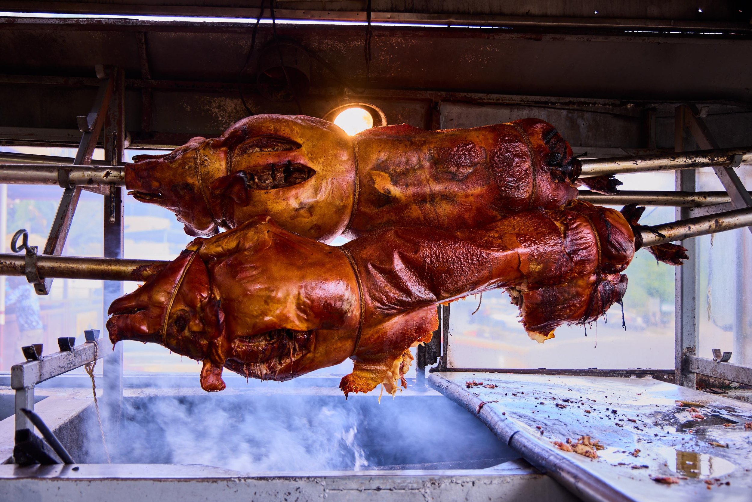 A roasted pig on a rotisserie spit in a commercial kitchen with smoke and heat rising below.