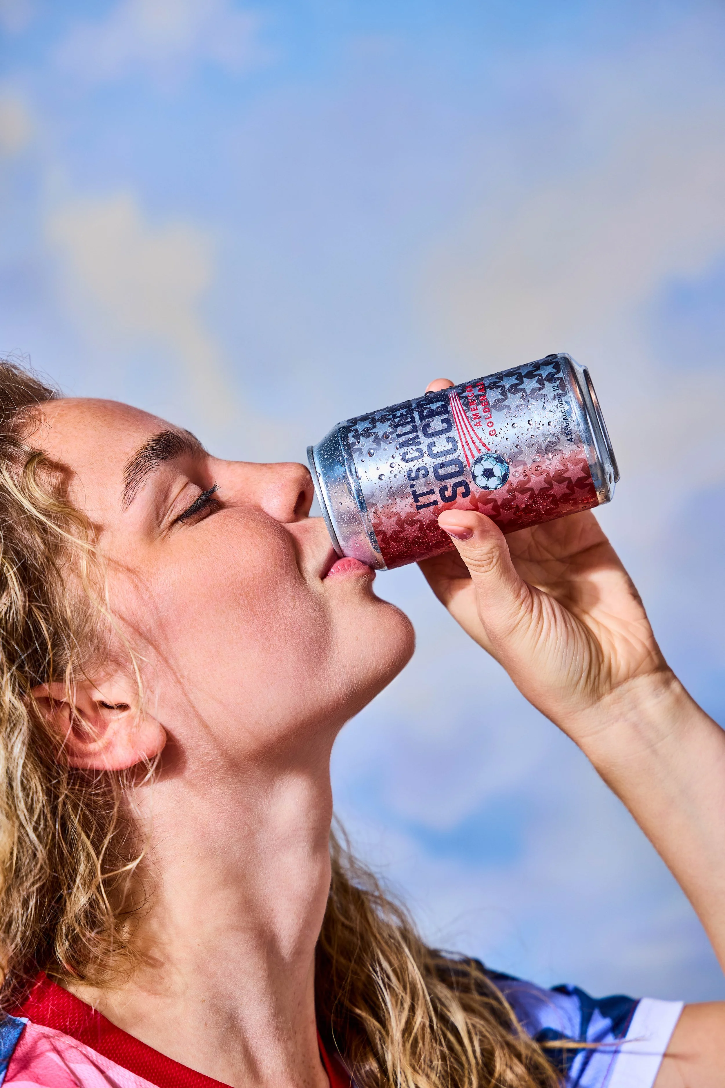 A woman with curly hair drinking from a cans of soda with patriotic branding, against a cloudy sky.