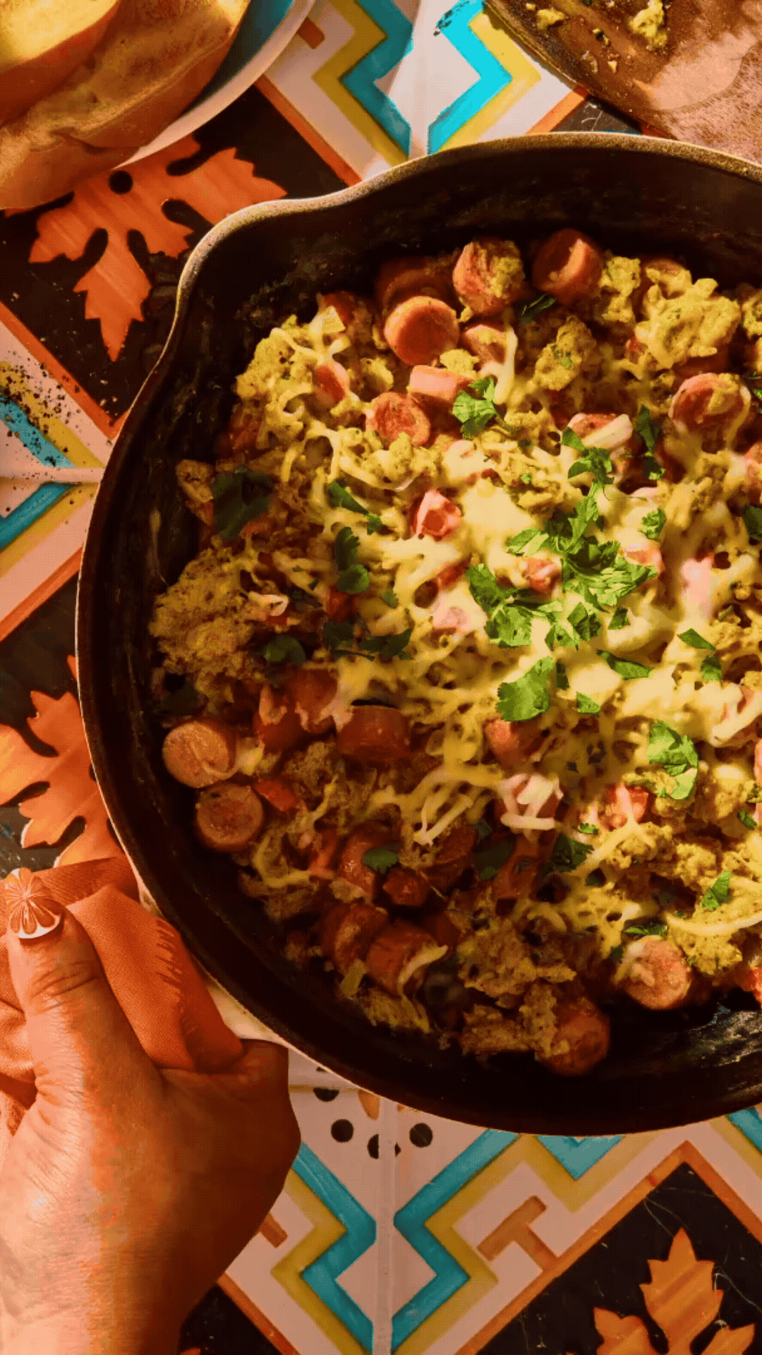 A hand holding a black skillet filled with cooked sausage, scrambled eggs, melted cheese, and chopped herbs, placed on a table with a colorful, geometric patterned tablecloth.