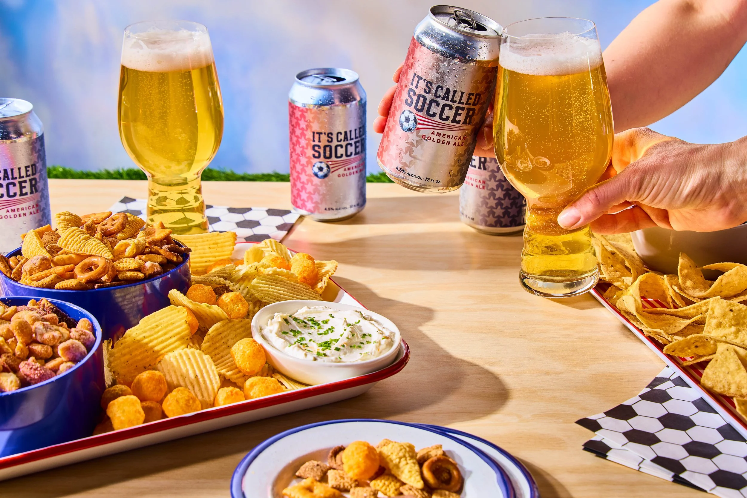 People celebrating with beers and snacks on a wooden table, including chips and dip, in a soccer-themed setting.