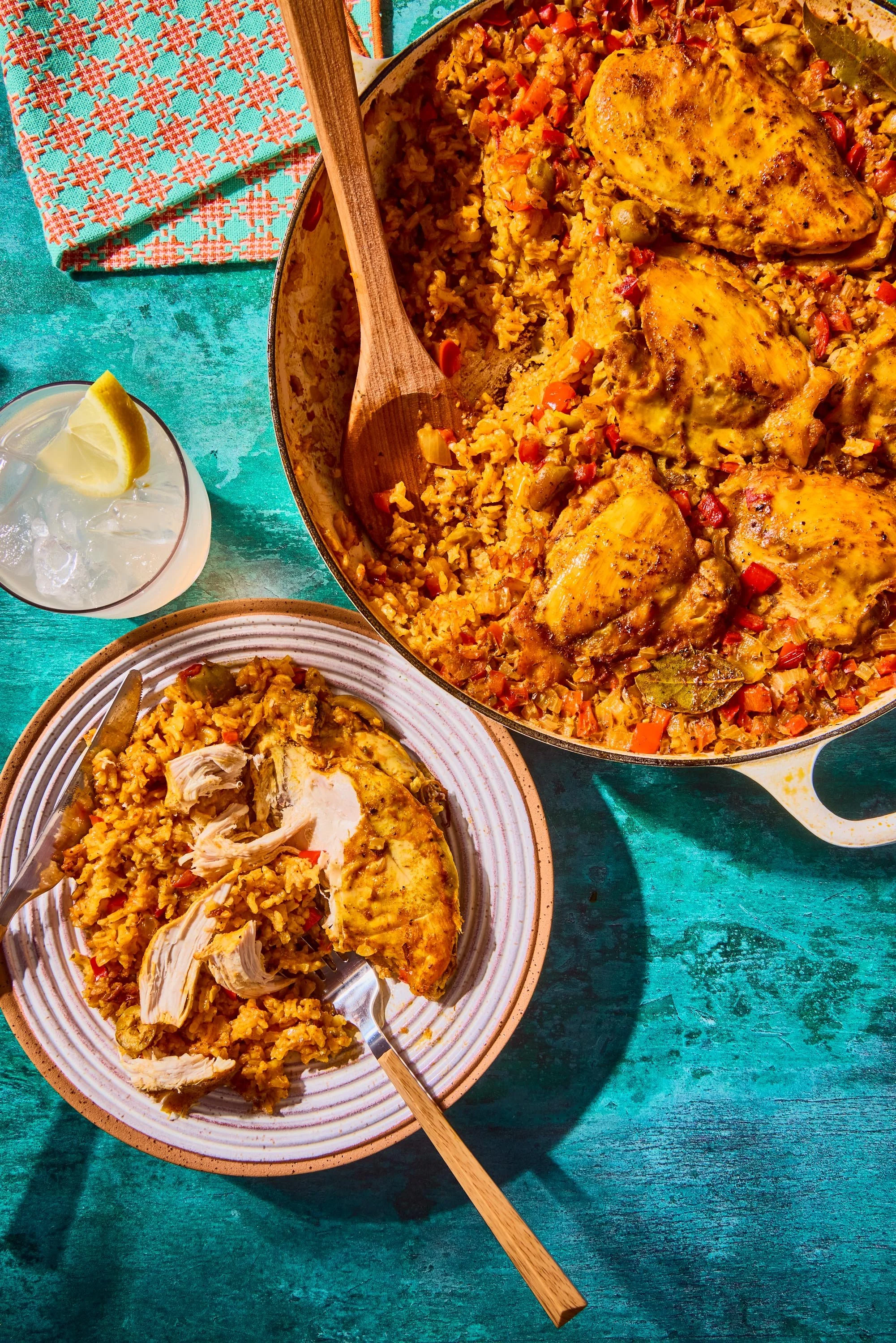A plate of cooked chicken with rice and vegetables, a glass of water with lemon, a colorful napkin, and a smaller bowl of rice on a teal wooden table.
