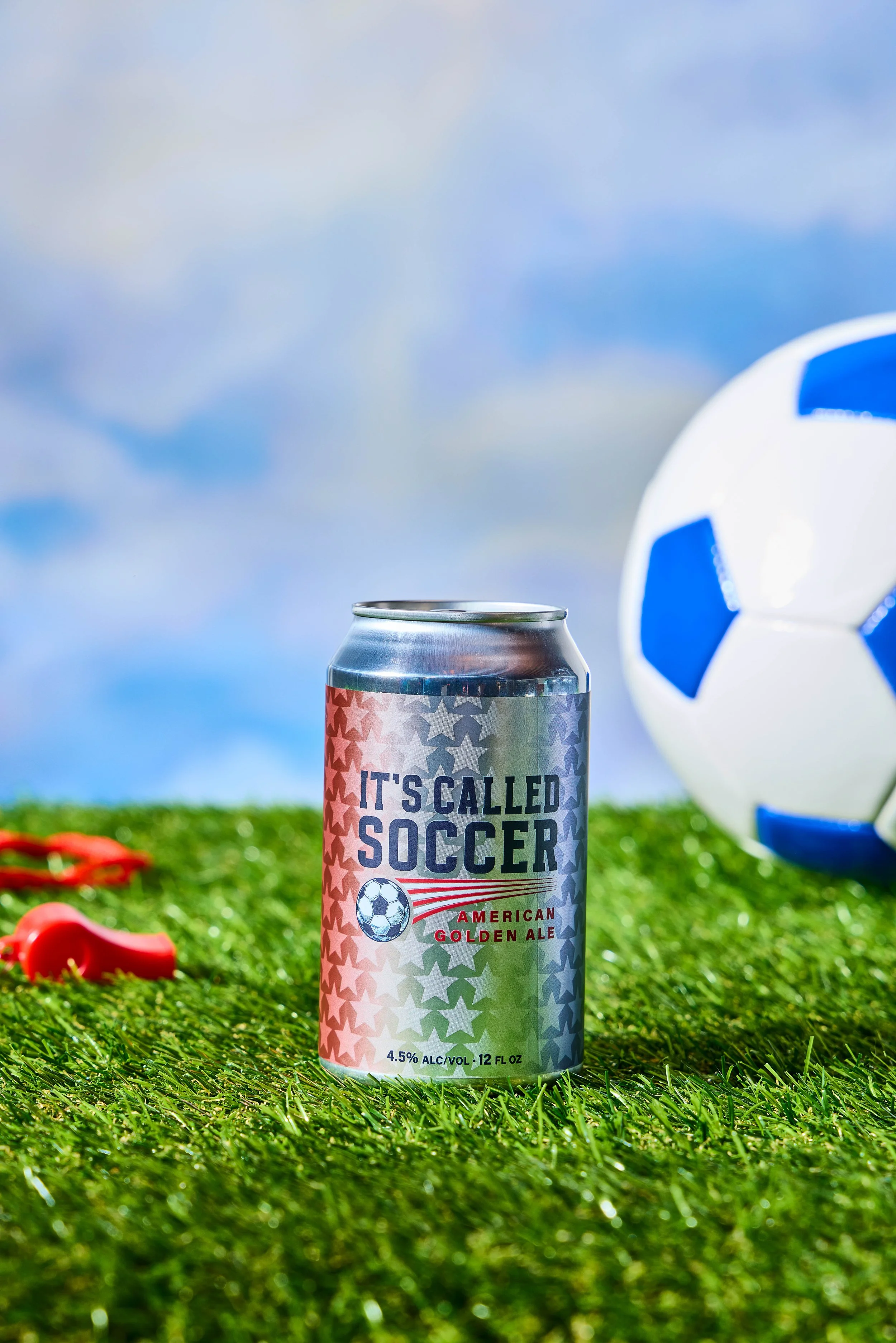 A can of beer labeled 'It's Called Soccer' with a soccer ball graphic, placed on a grassy field next to a soccer ball and red whistle, against a partly cloudy sky.