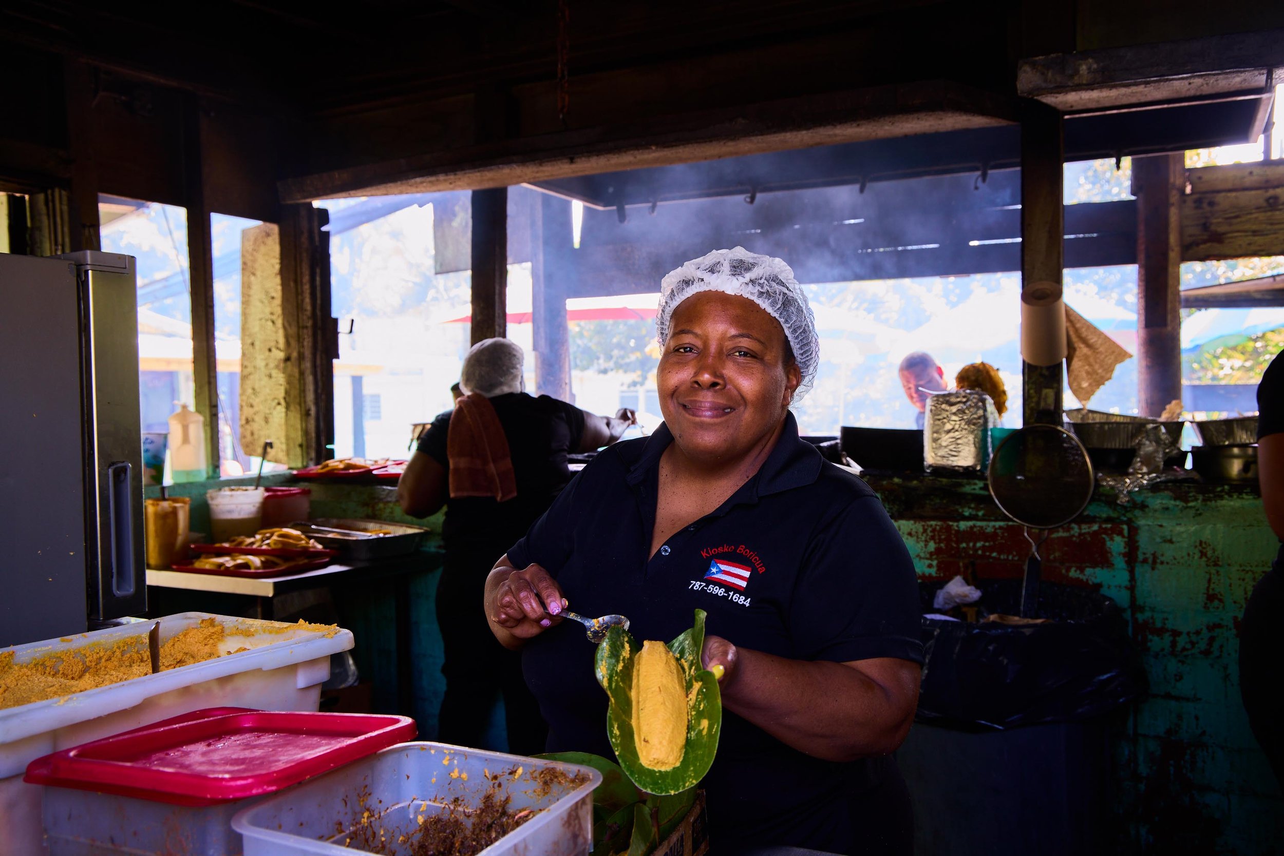 A woman working at a food stand, wearing a hairnet, with containers of food and ingredients on the counter, in a rustic open-air setting.