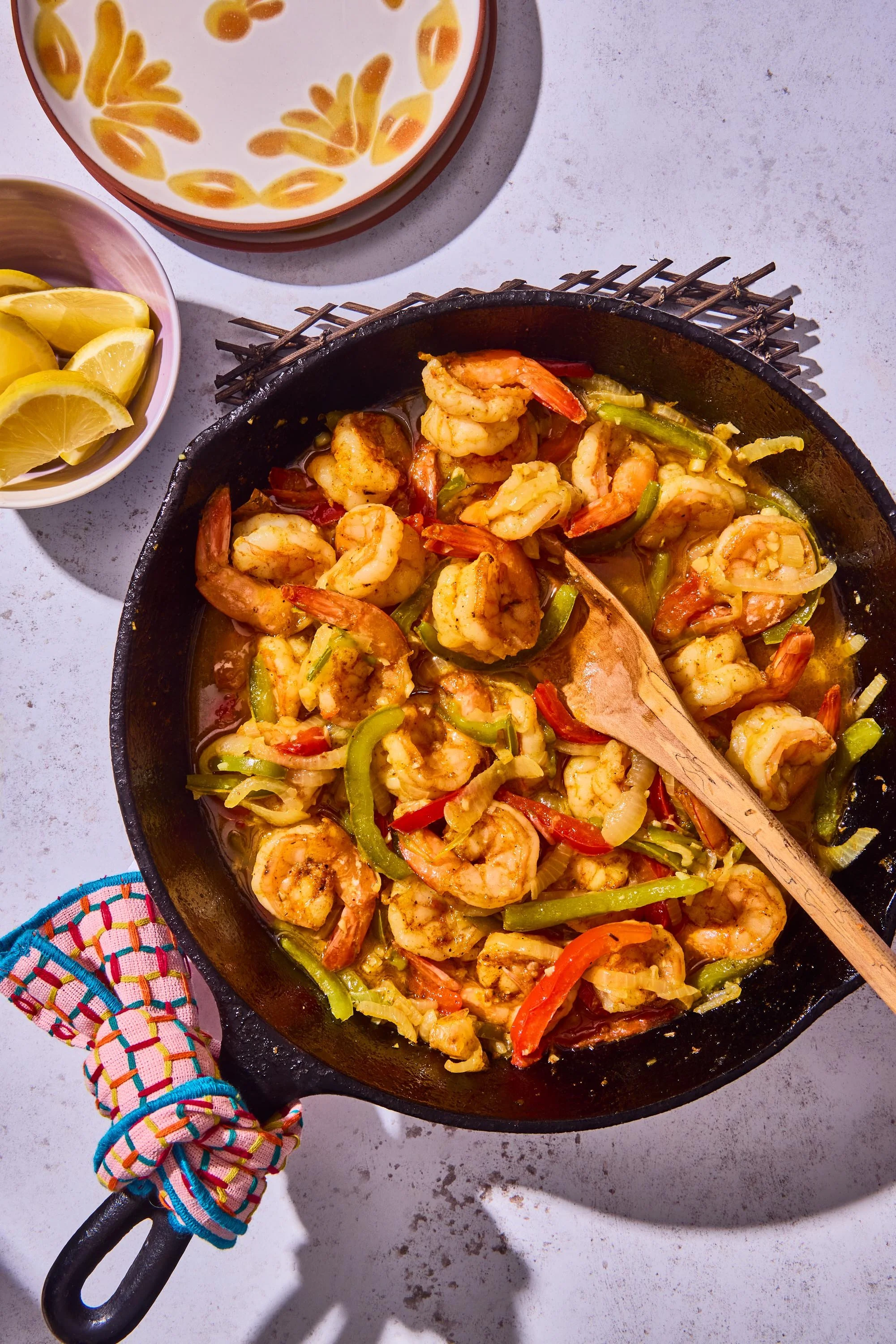 Shrimp stir-fry with red and green bell peppers cooking in a black skillet. Lemon wedges in a white bowl and another bowl with a yellow-orange floral pattern are nearby. A colorful woven cloth is partially visible on the lower left corner.