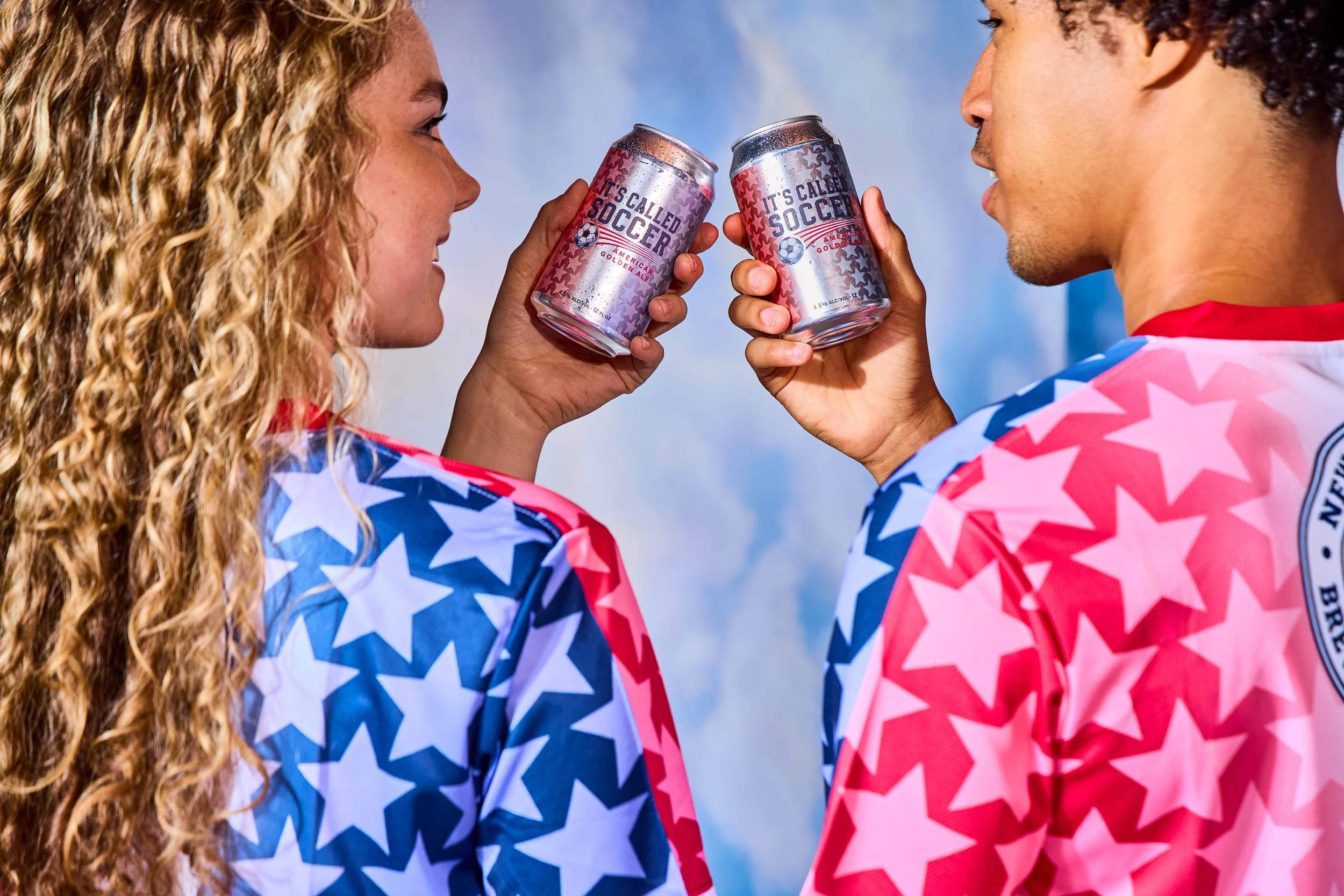 A man and woman, dressed in red, white, and blue star-patterned sports jerseys, are facing each other and toasting with cans of beer against a cloudy sky.