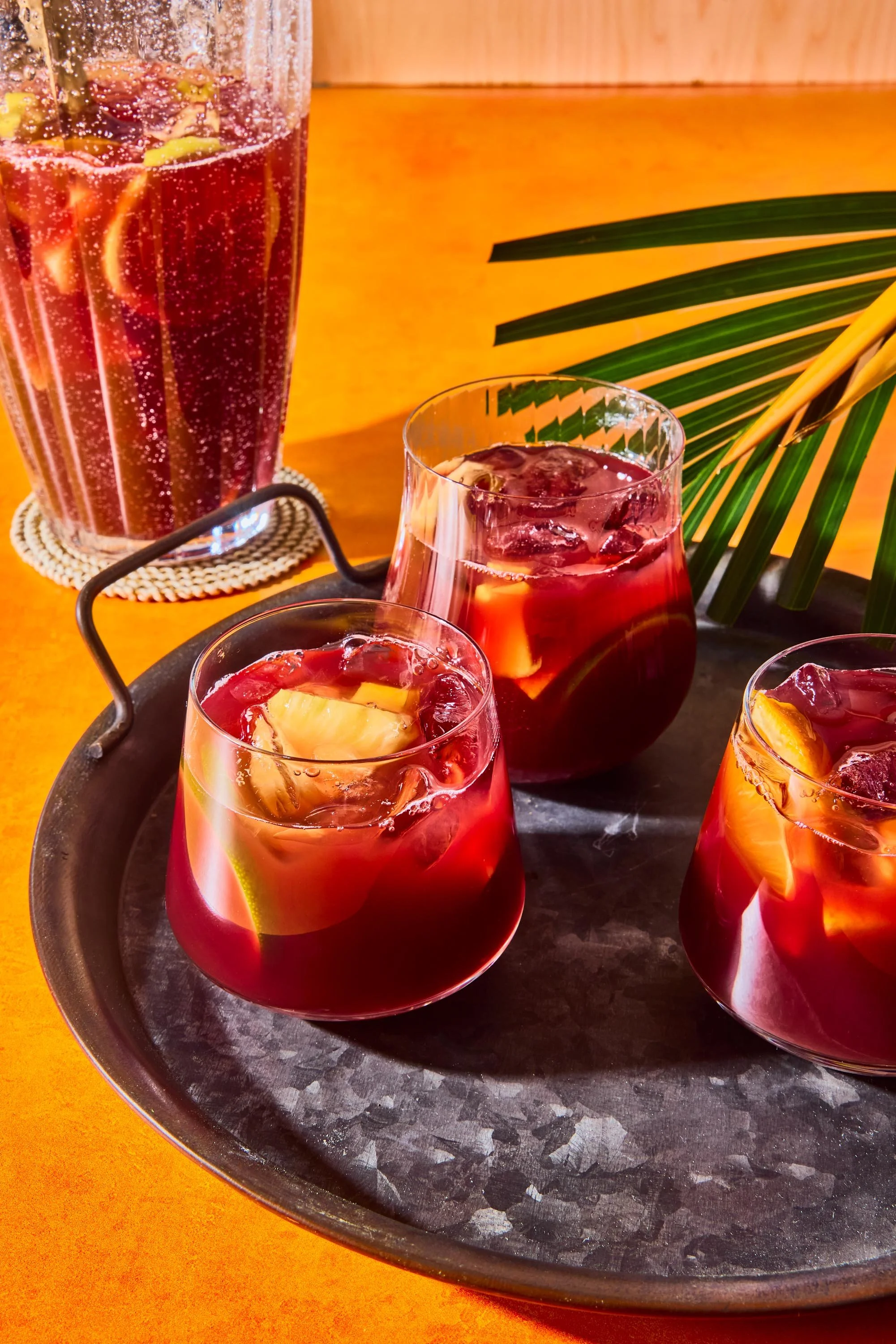 Glass pitcher and glasses filled with a dark red beverage, garnished with fruit slices, on a black tray with a palm leaf prop in the background.
