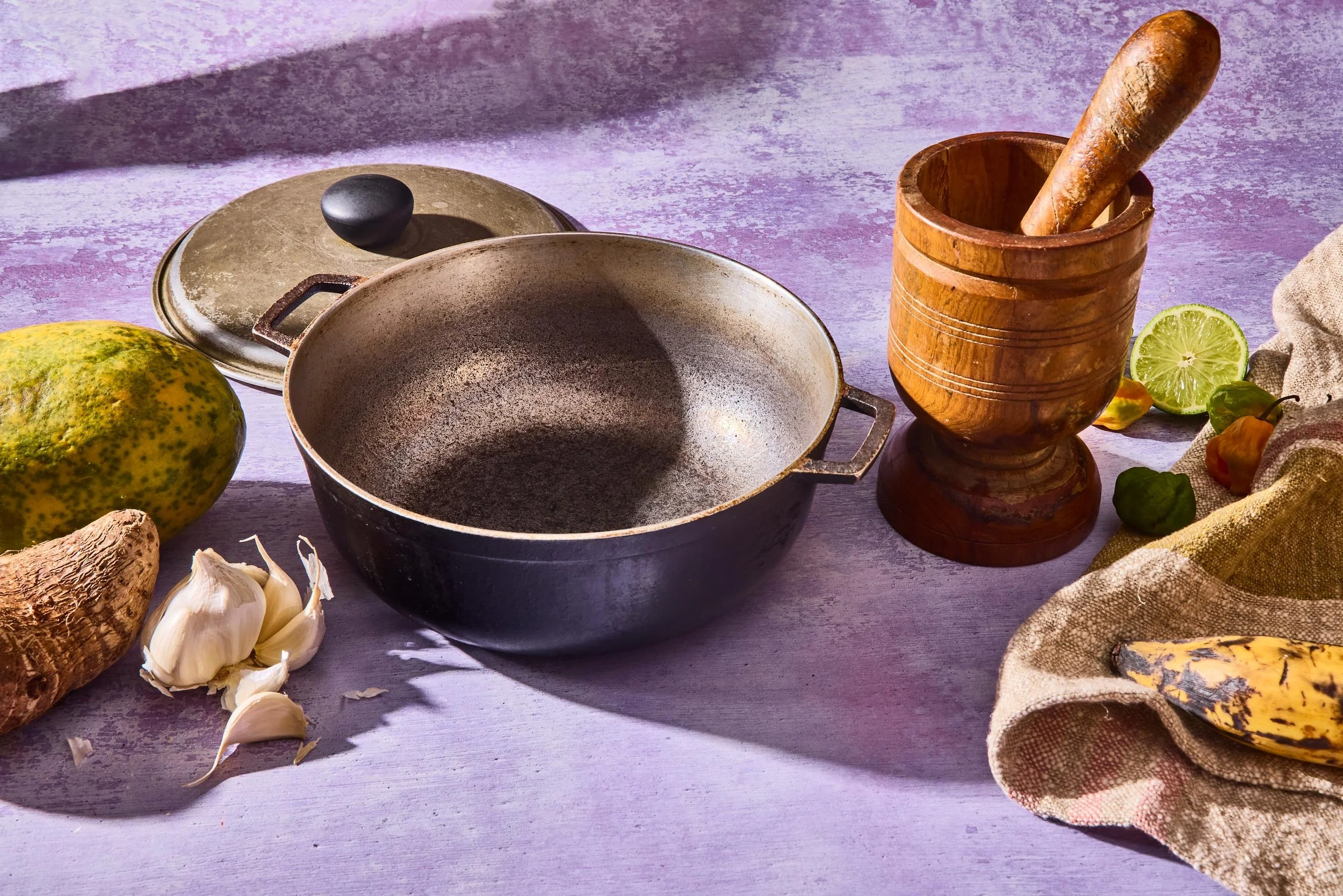 A wooden mortar and pestle, a large mixing bowl with a handle, garlic cloves, and various vegetables including a lemon, a small green and yellow pepper, and a yellow gourd on a purple-textured surface.