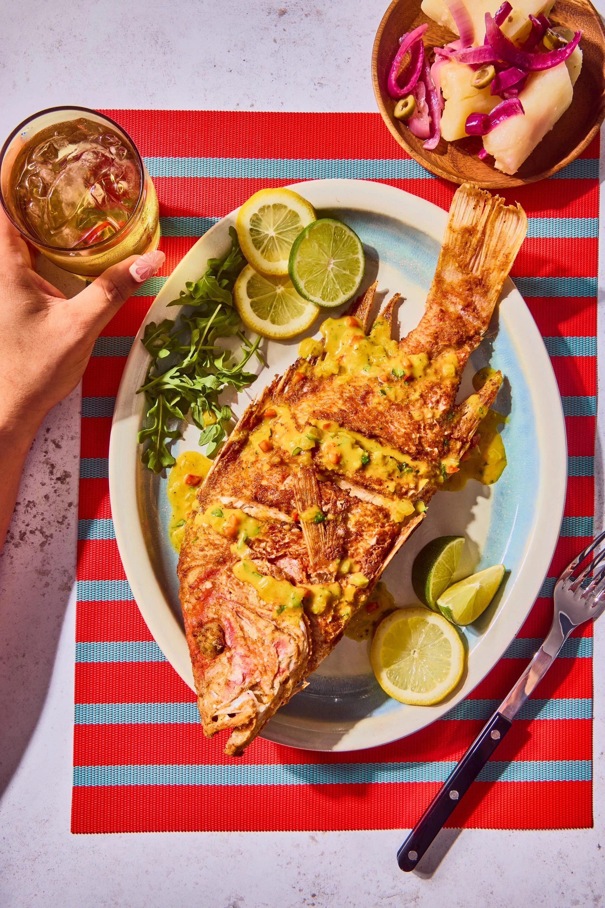 A whole grilled fish topped with a yellow sauce, garnished with lime slices, served on a white plate with lemon wedges and greens. A glass of a beverage with ice is held by a person with a painted nail. A side dish of boiled potatoes with pickled oni