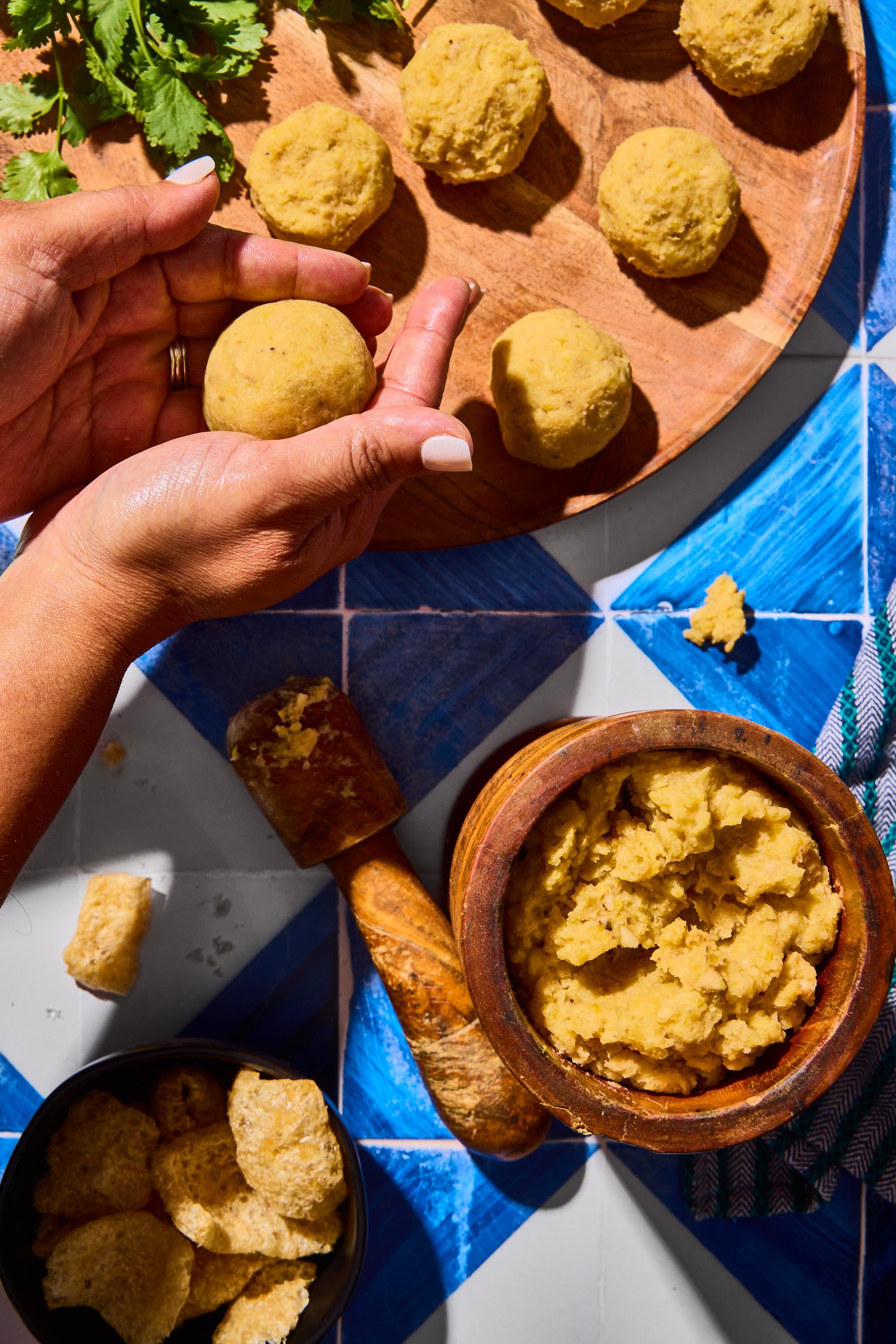 A person holding a ball of yellow masa dough, with several other balls of masa on a wooden cutting board. There are also bowls with more masa, chunks of fried pork rinds, and a blue and white tiled surface.