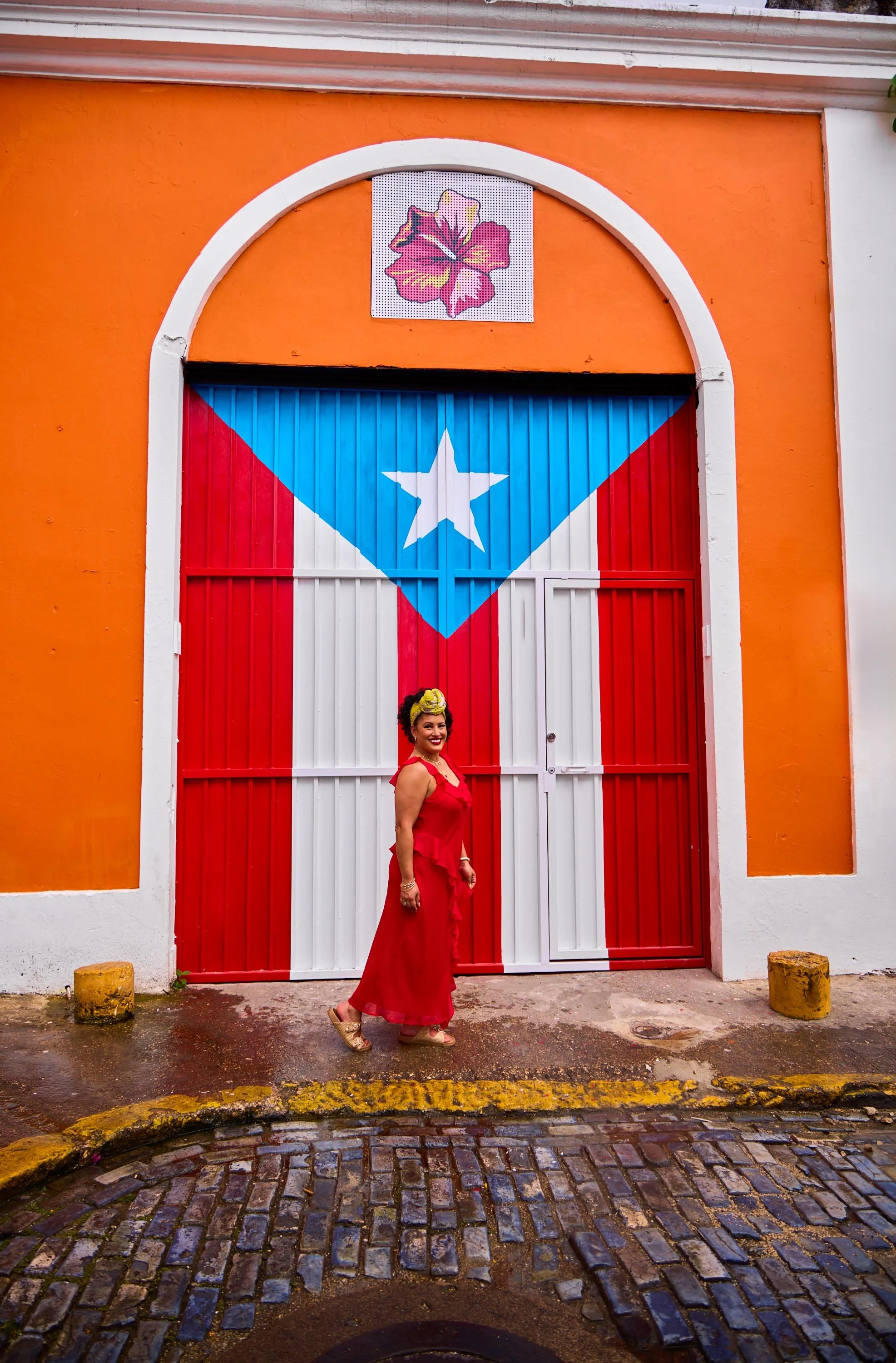 A woman in a red dress and yellow headscarf standing in front of a bus door painted with the Puerto Rican flag and a colorful flower sign above the door, on a cobblestone street.